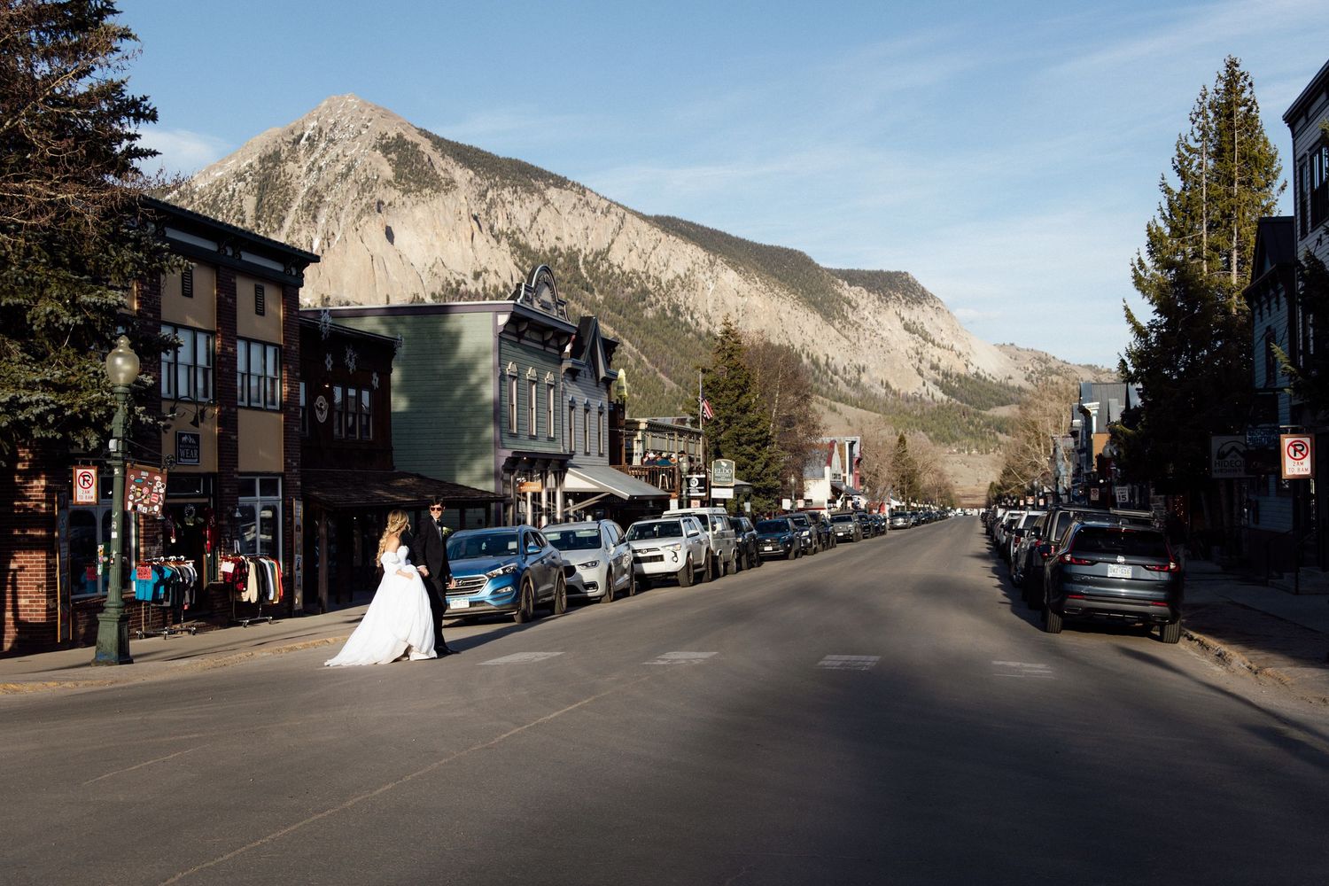Bride and groom cross a mountain town street in Crested Butte, Colorado, with stunning rocky peaks in the background.