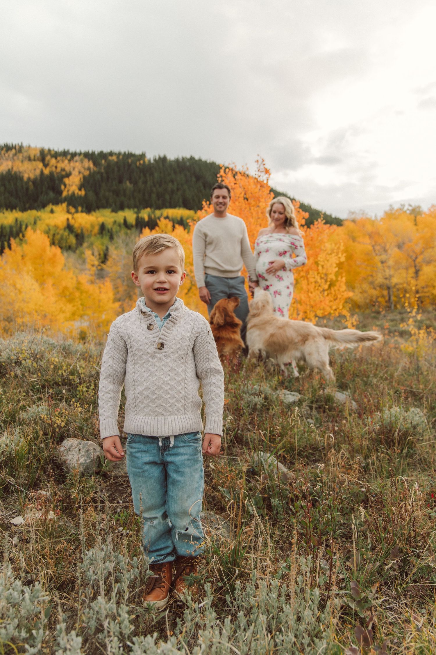Young boy in knit sweater stands in autumn field with parents, golden retriever, and colorful fall foliage backdrop.