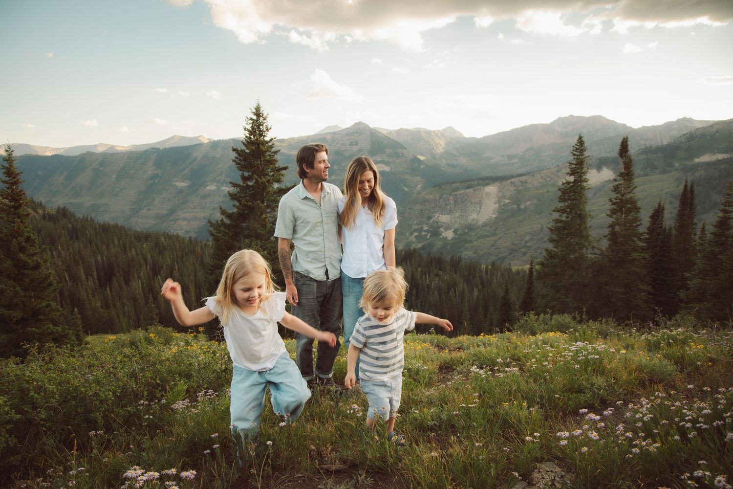 Family of four enjoying a scenic mountain meadow with evergreen trees and peaks in the background on a sunny day.