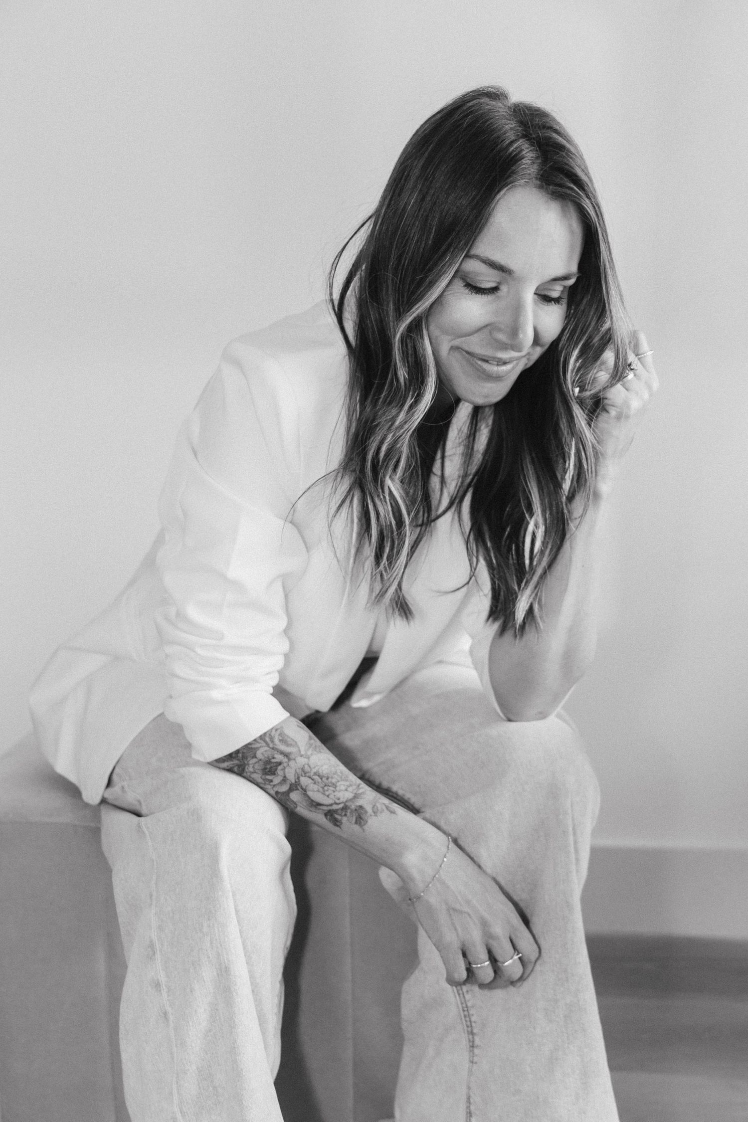Black and white portrait of a smiling woman with long wavy hair and a forearm tattoo, seated in a white blazer and wide-leg pants.