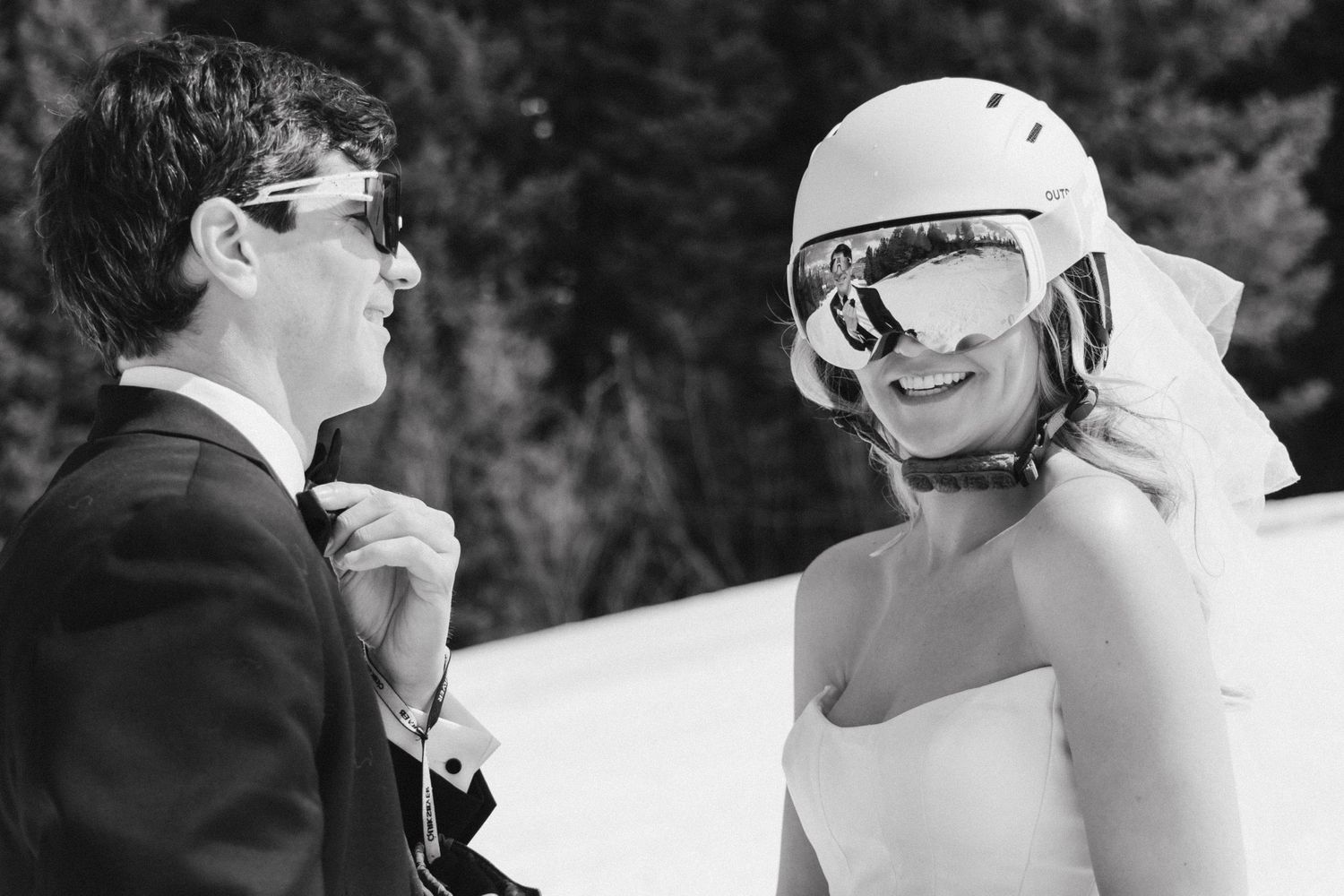 Black and white photo of a smiling bride in a white strapless gown and ski helmet with goggles beside a groom in a suit and sunglasses.