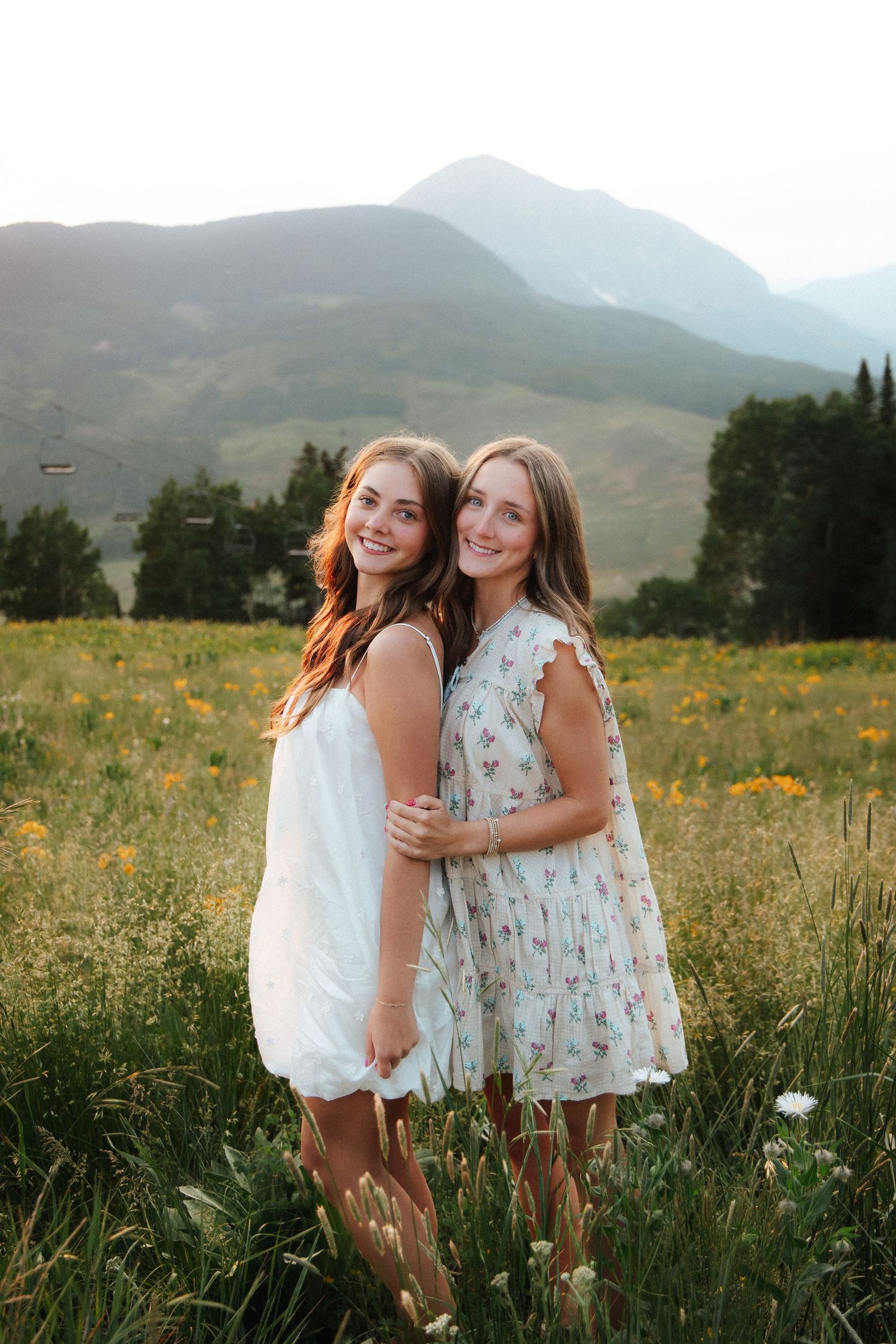 Two smiling young women in summer dresses pose in a wildflower meadow with mountains in the background.