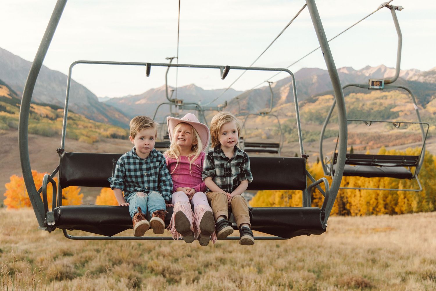 Three children sit together on a ski resort chairlift with autumn mountain scenery and golden trees in the background.
