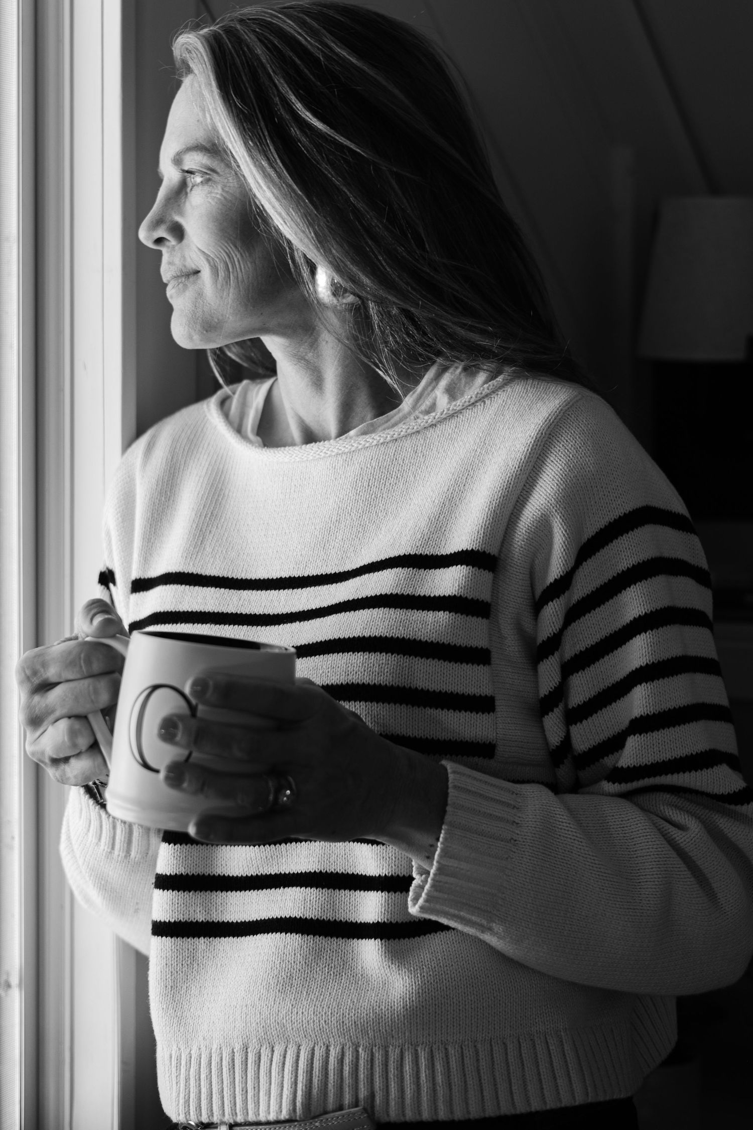 Black and white photo of someone in a striped sweater holding a coffee cup while looking out a window.
