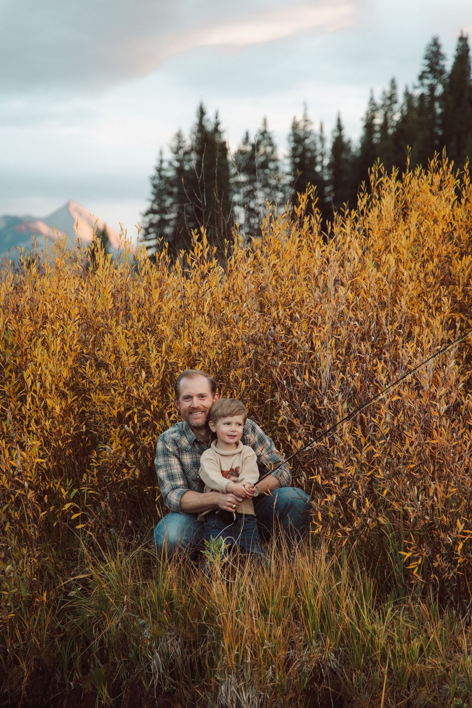 Two people sit together surrounded by golden autumn brush and evergreen trees in a mountain landscape at dusk.