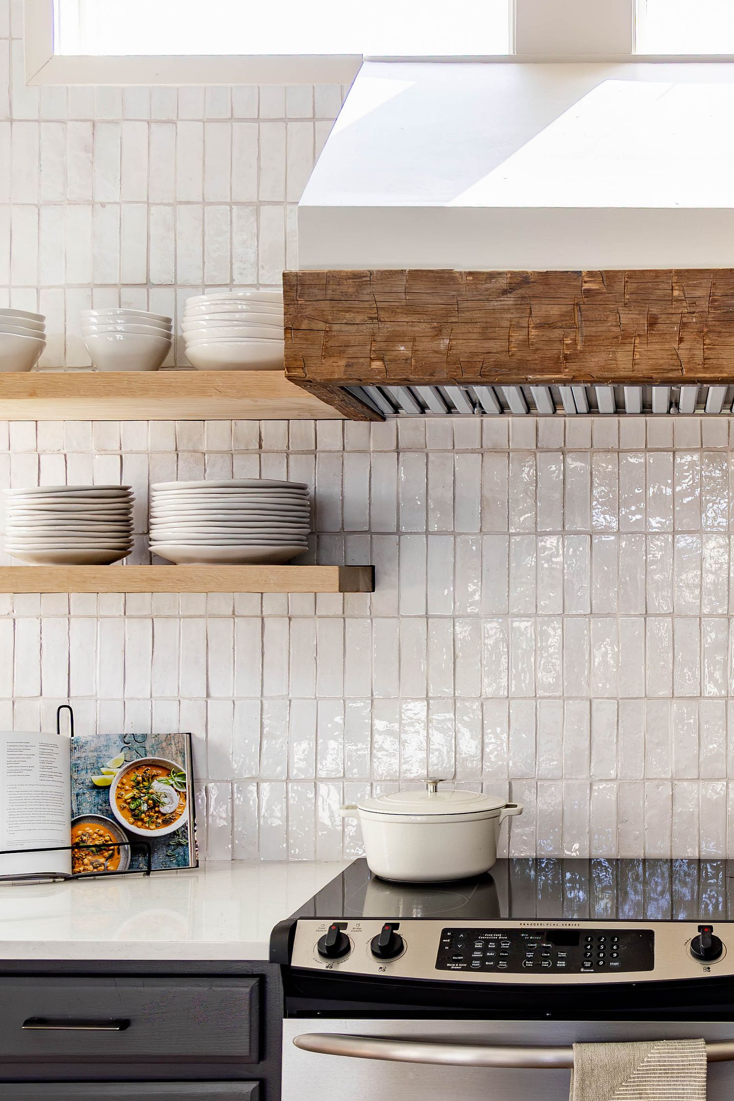 Modern kitchen with white zellige tile backsplash, wood open shelves holding stacked dishes, and a stainless steel range with dutch oven.