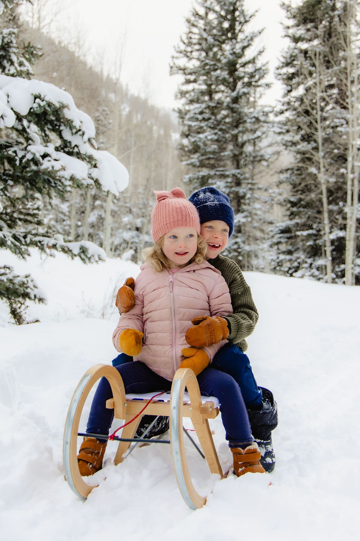 Two bundled-up children share a wooden sled on a snowy winter trail surrounded by evergreen trees.