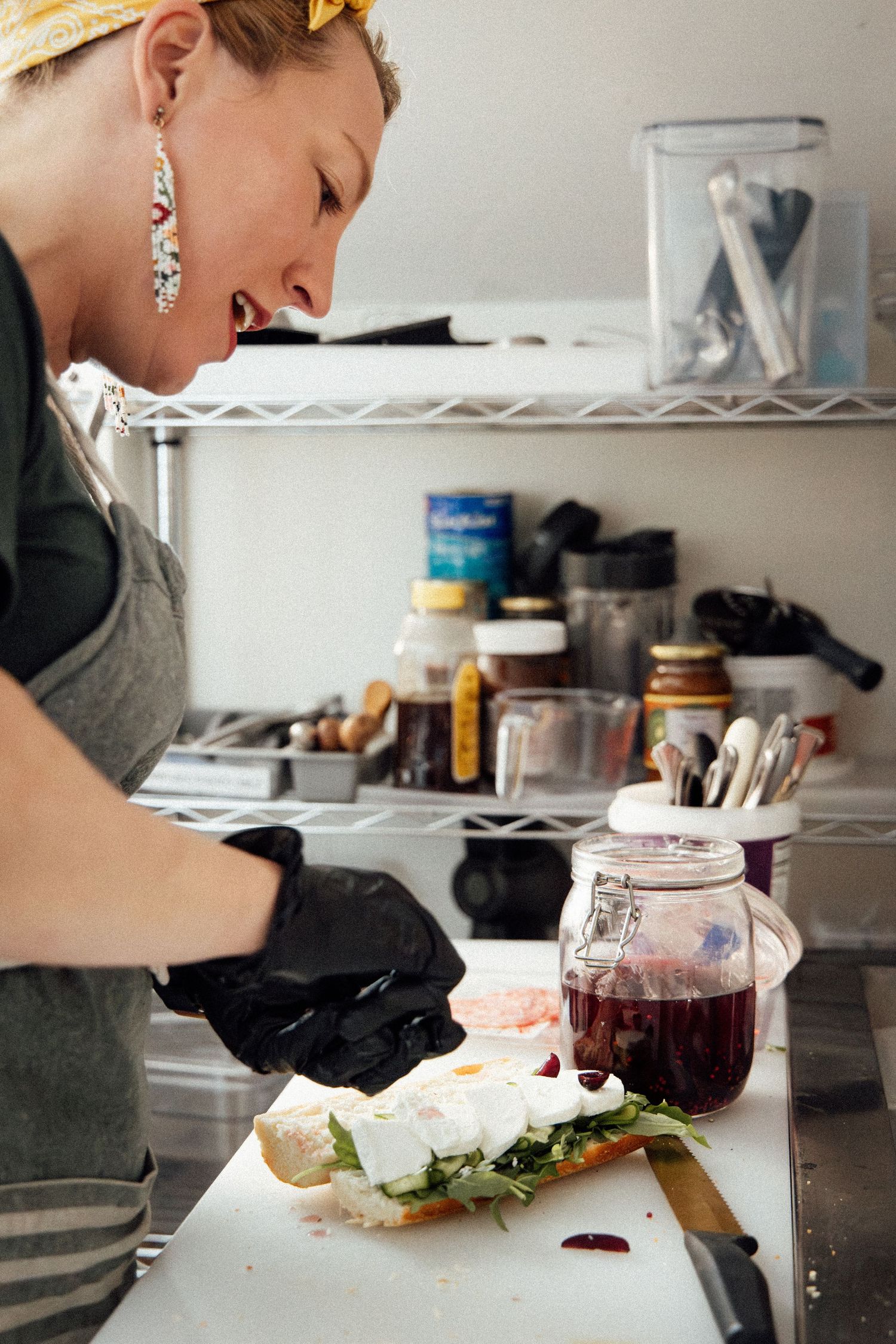 Woman in black gloves preparing a gourmet sandwich on a white cutting board in a professional kitchen.