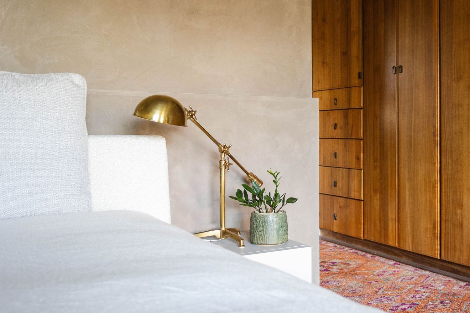 Cozy bedroom corner with brass desk lamp, small plant, white bedding, and wooden dresser on a vintage rug.