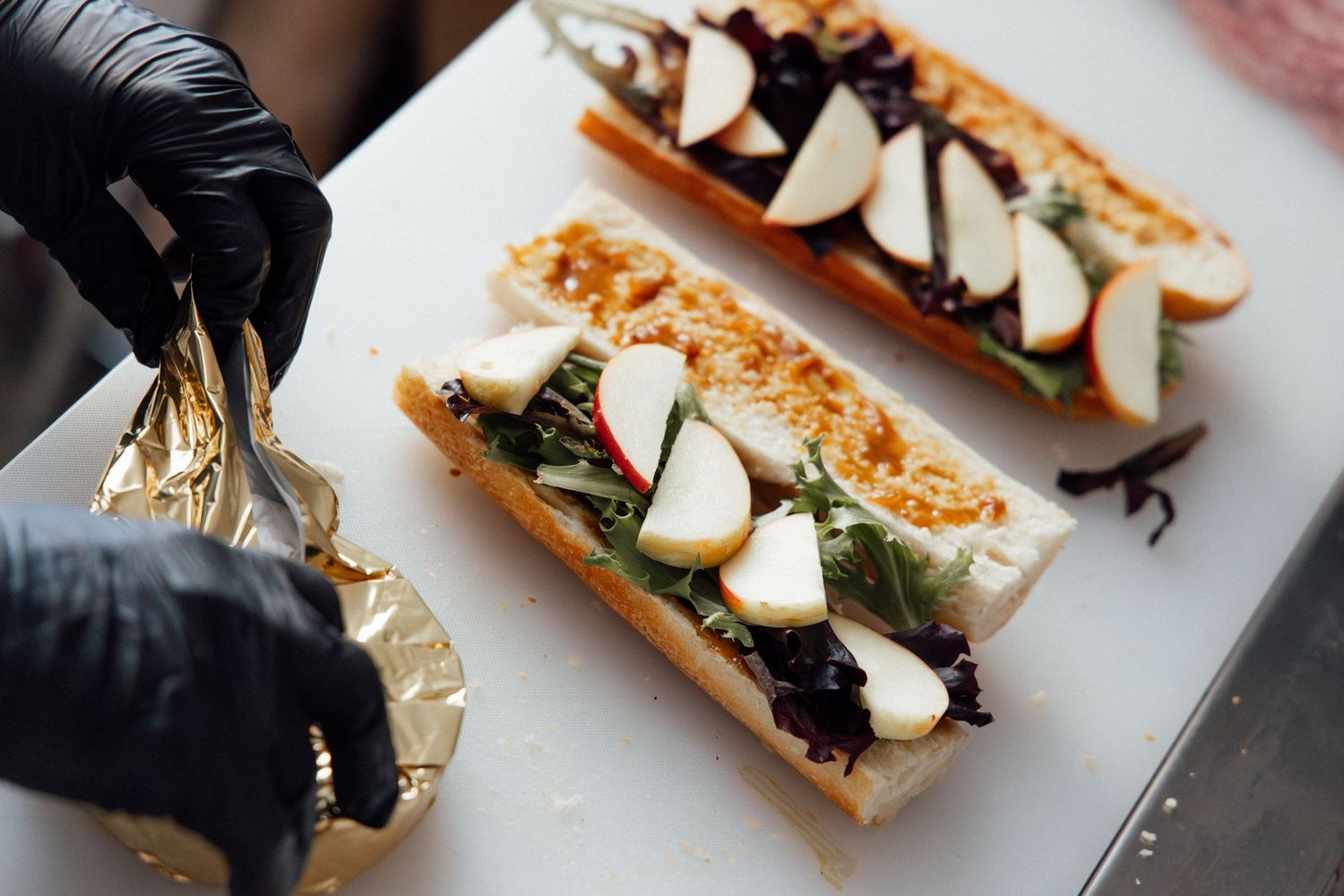 Chef in black gloves assembling gourmet sandwiches topped with apple slices and greens on a white cutting board.