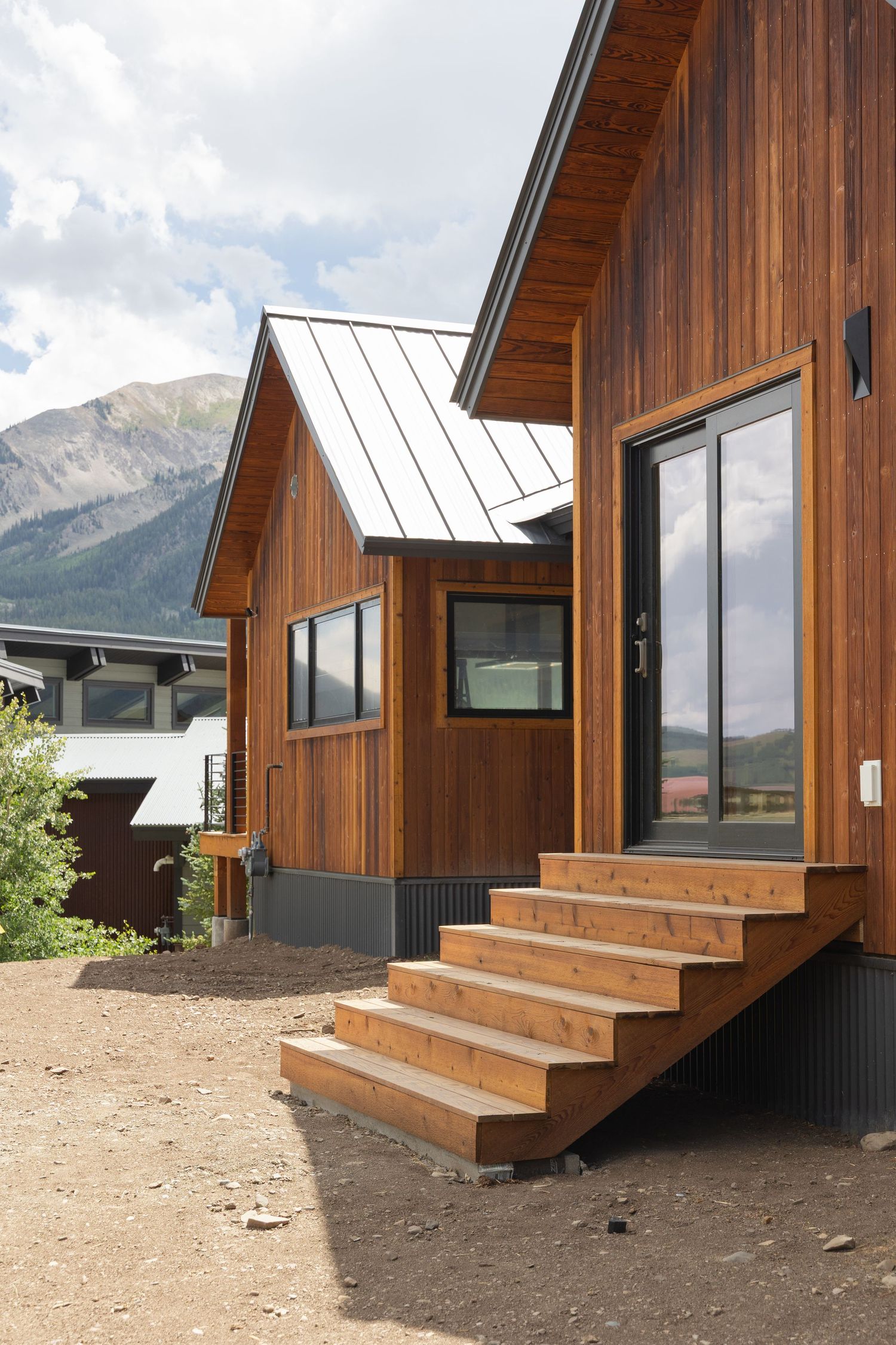 Modern wood-clad mountain cabin with cedar siding, metal roof, wooden steps, and black-framed windows against Rocky Mountain backdrop.