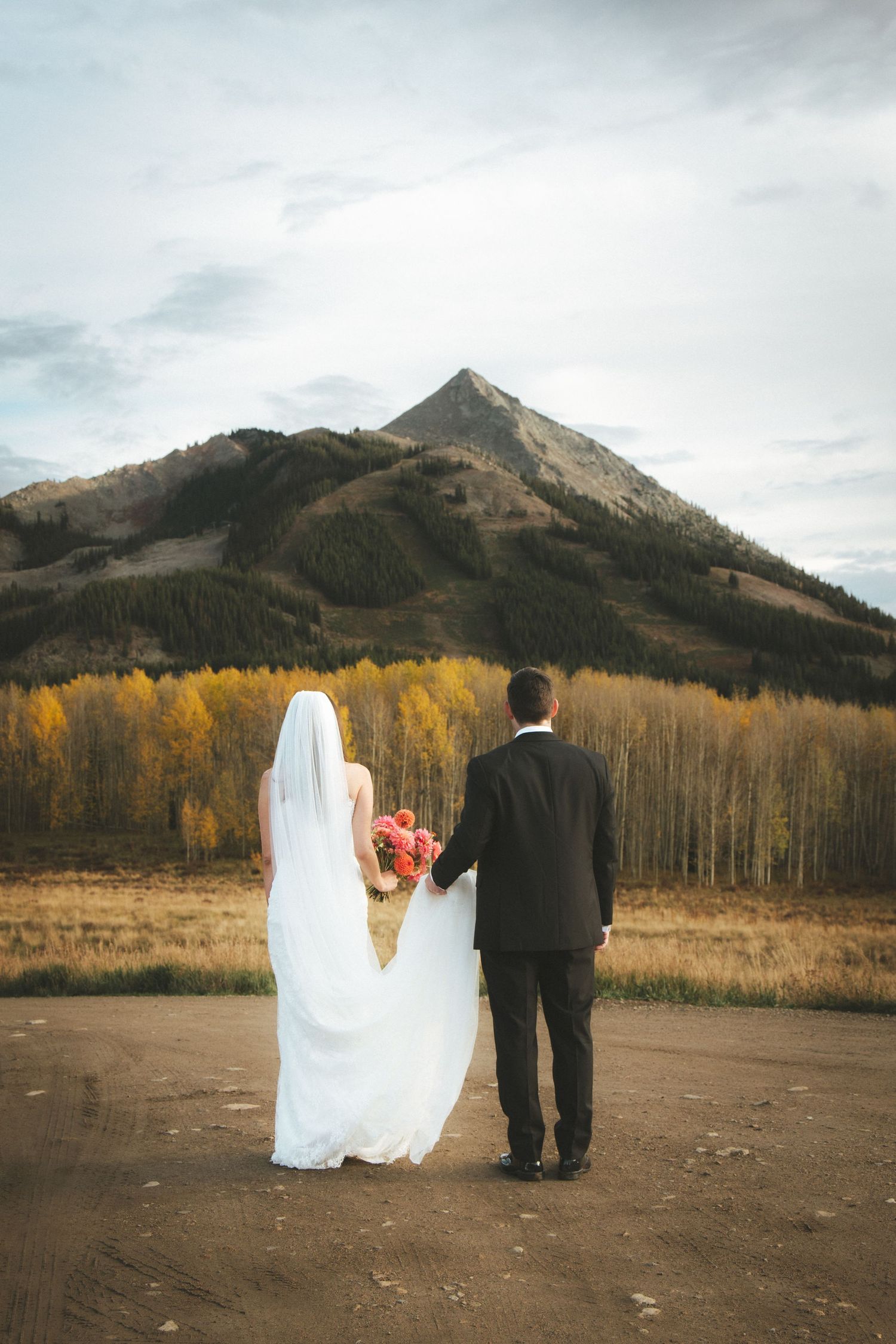 Bride in a white gown and veil holding pink flowers stands hand in hand with a groom before golden aspens and a mountain peak.