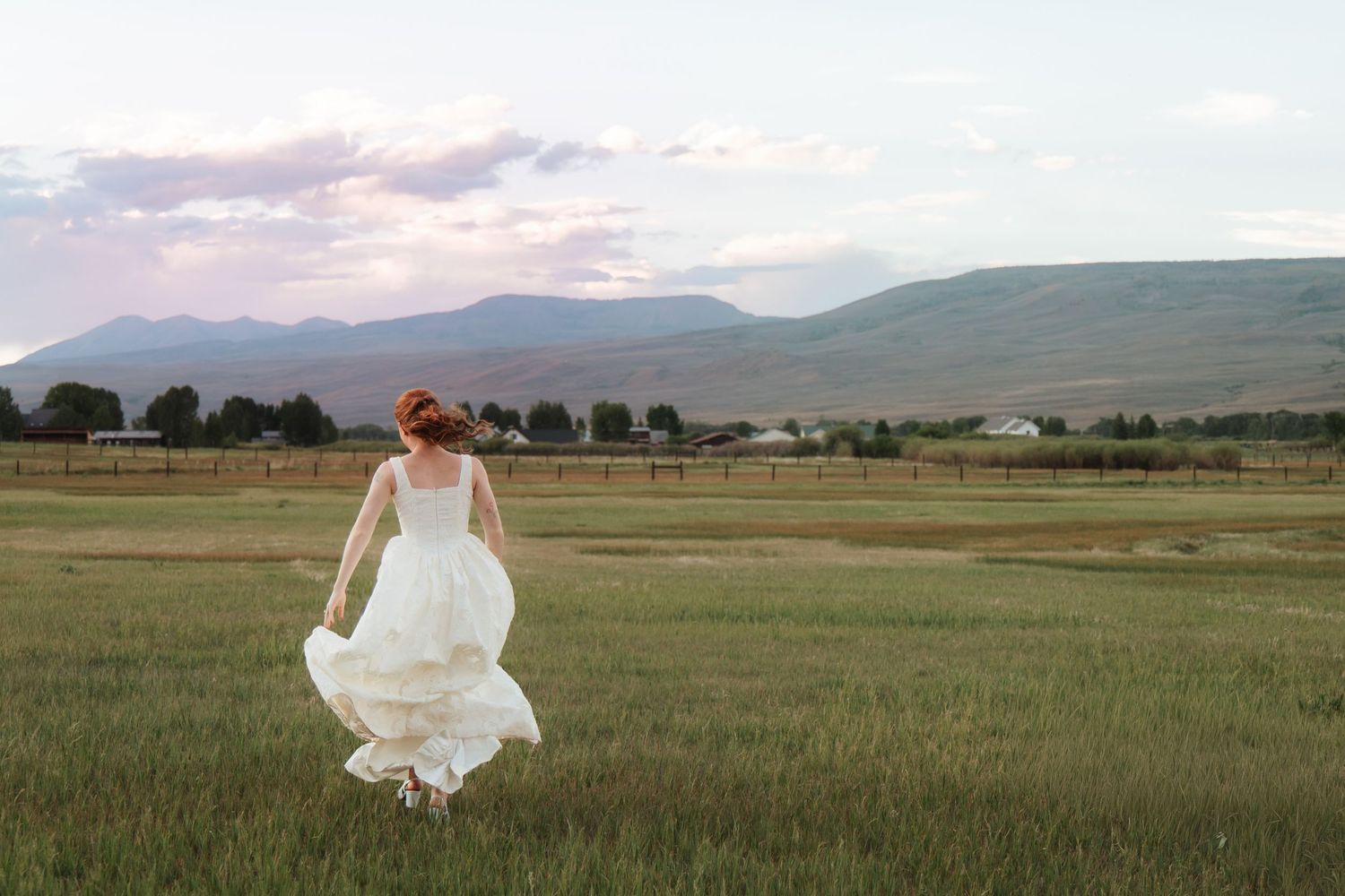 Bride in a flowing white wedding dress runs through a green grassy field with mountains and a pastel sunset sky in the background.