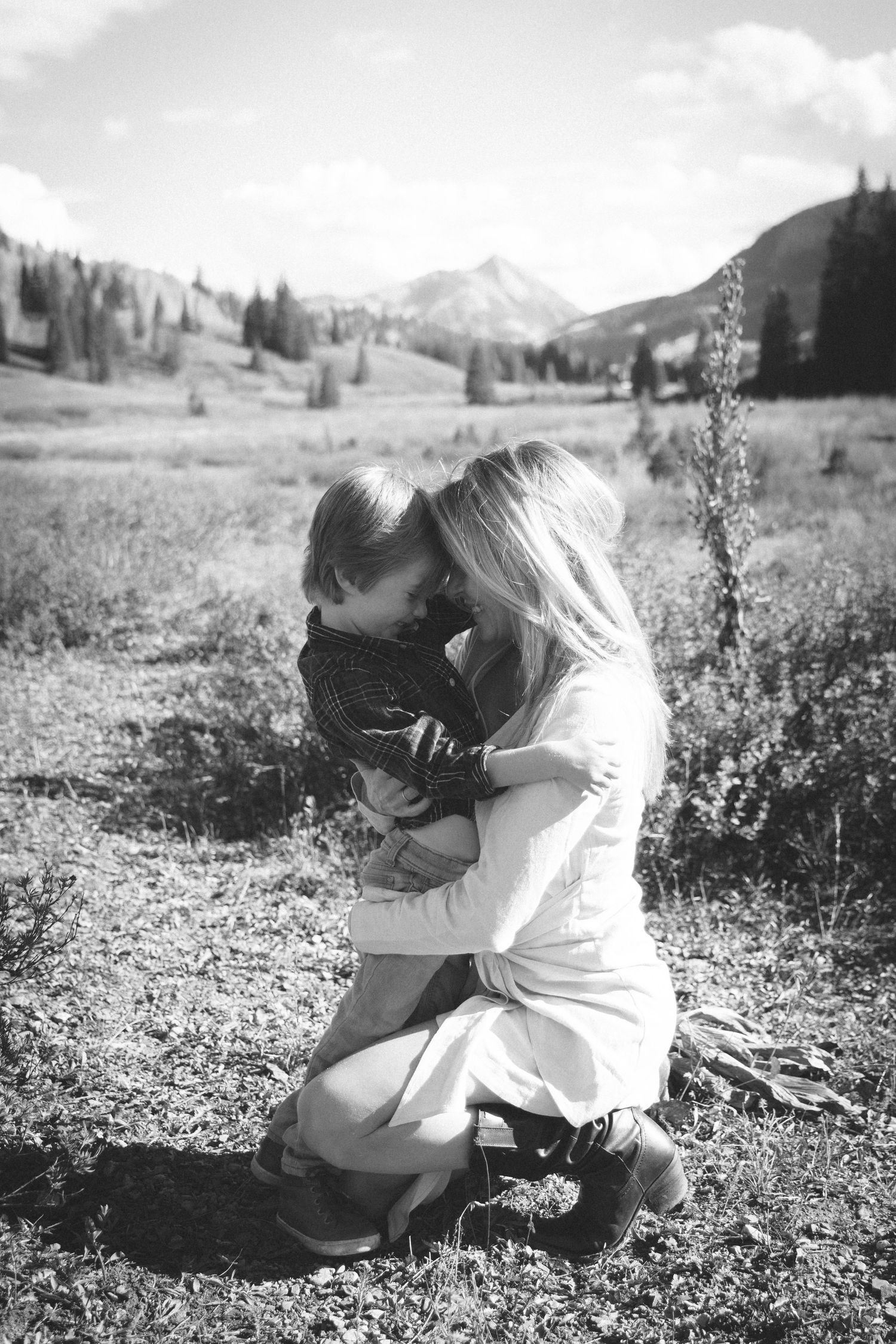 Black and white photo of a woman kneeling in a mountain meadow and embracing a young boy, with peaks and evergreen trees behind them.