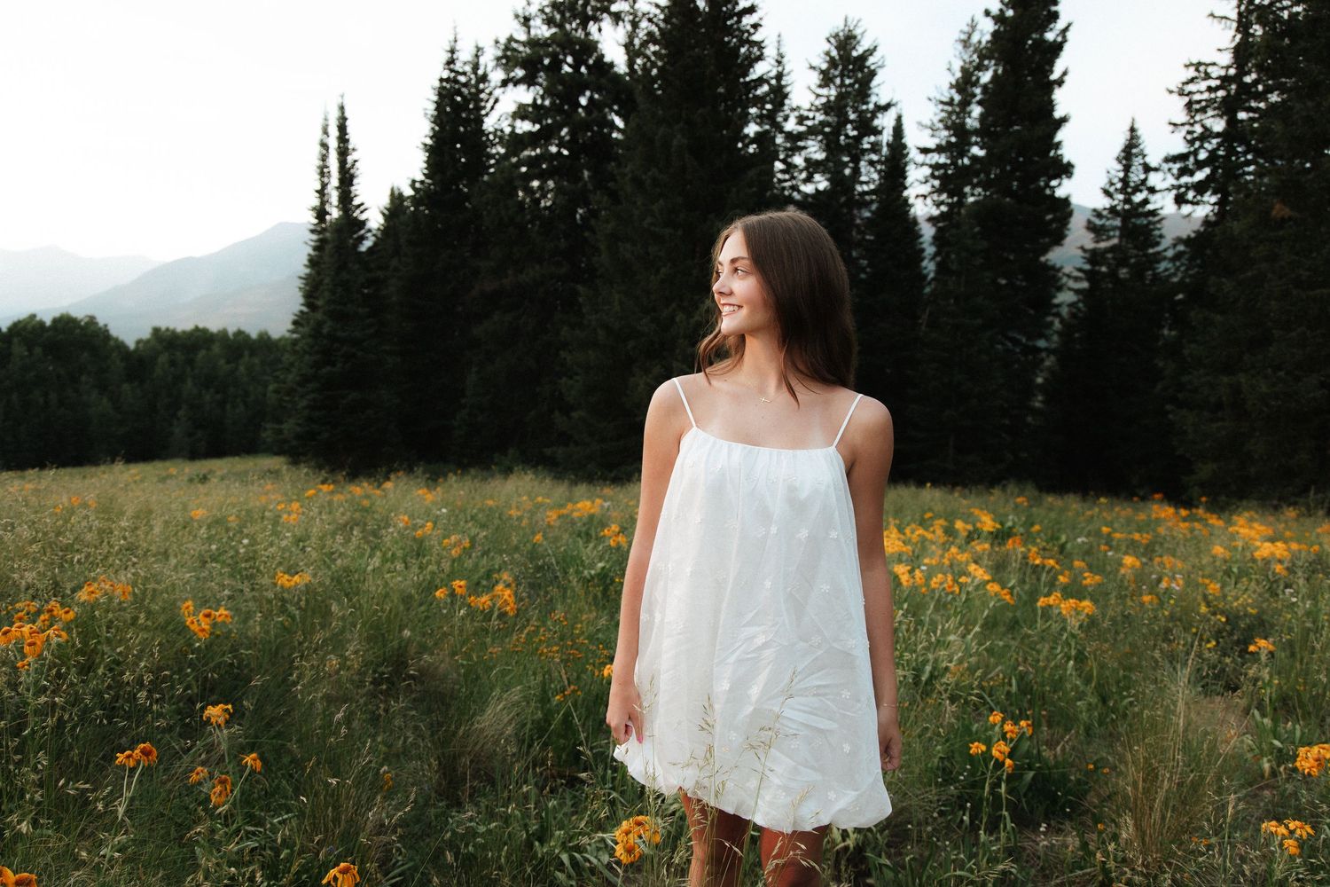 Young woman in white sundress standing in wildflower meadow with evergreen trees and mountains in background.