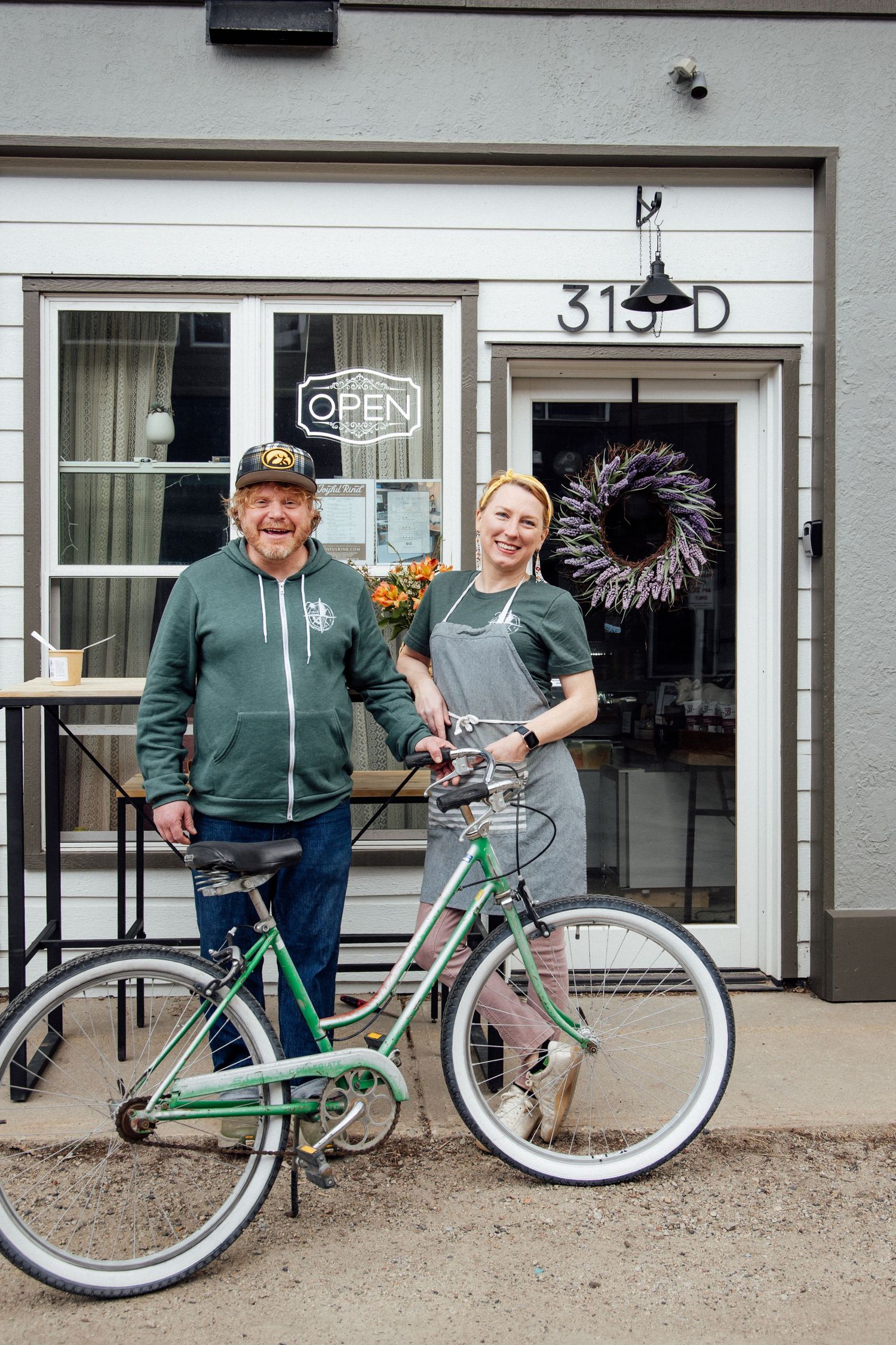 Two smiling people pose with a green bicycle outside an open shop at 313-D with a decorative wreath on the window.