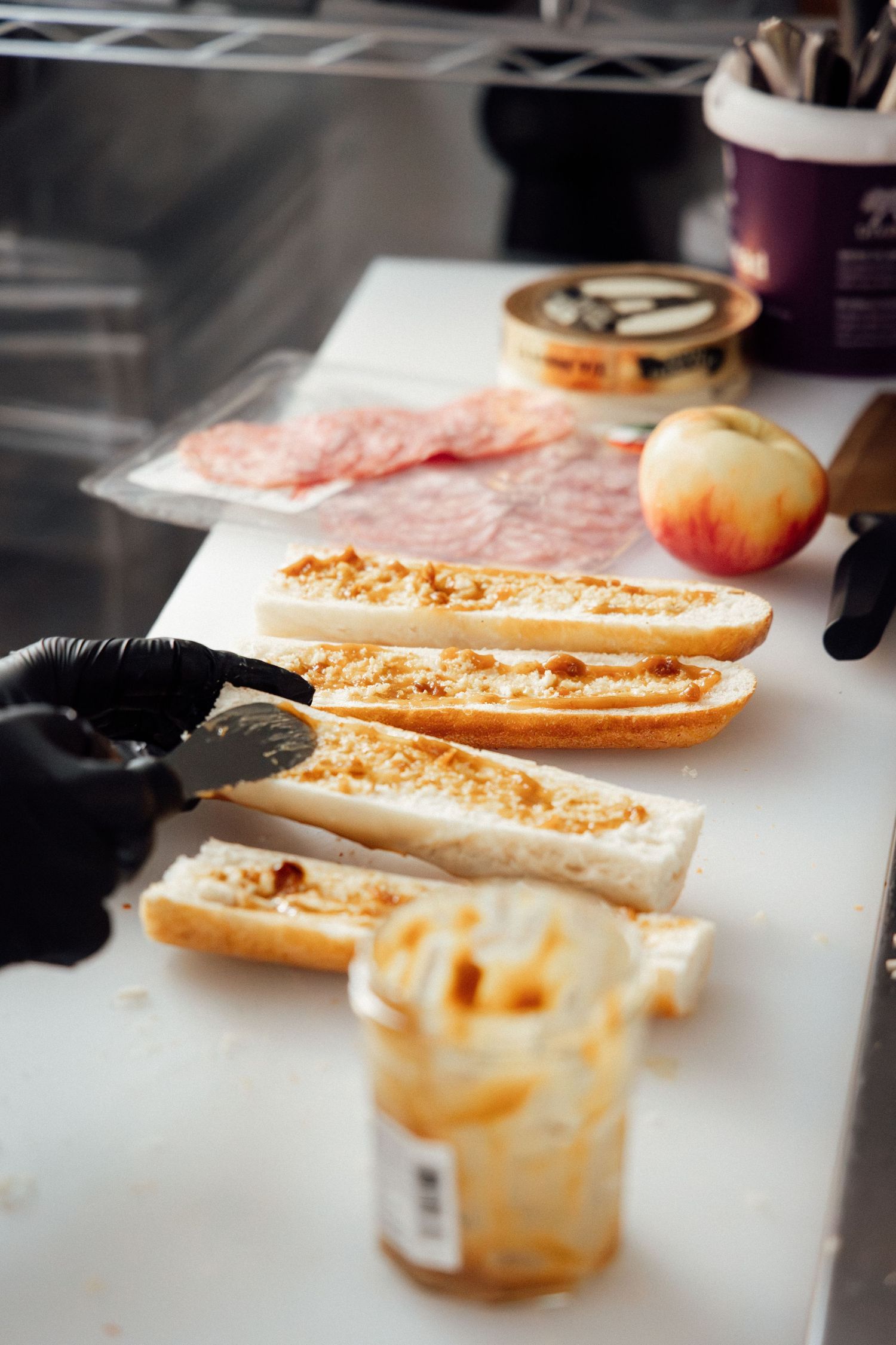 Chef in black gloves spreading jam on baguette halves with deli meat and apple on white cutting board.