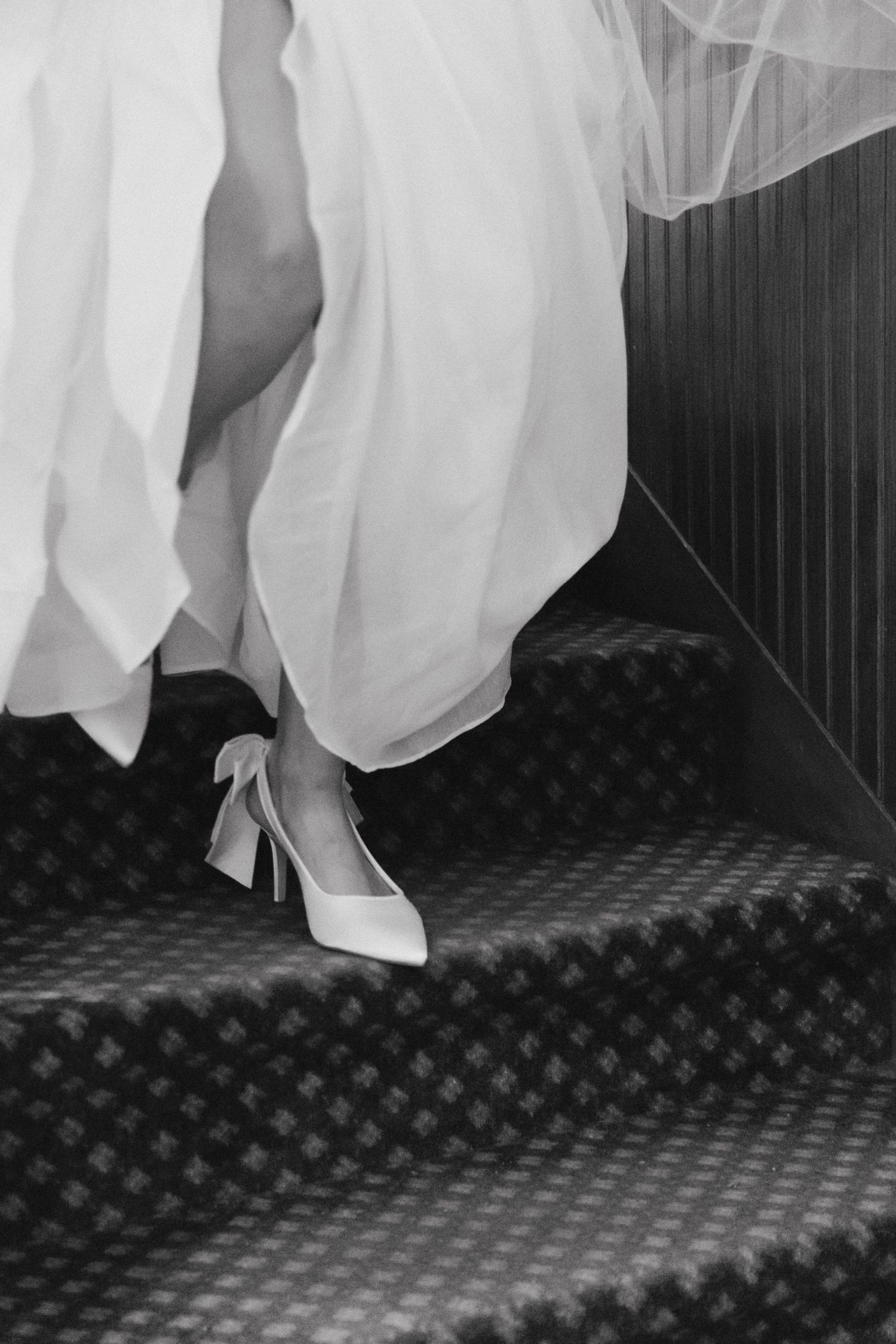 Black and white photo of a bride ascending stairs in elegant heels and flowing wedding dress with veil.