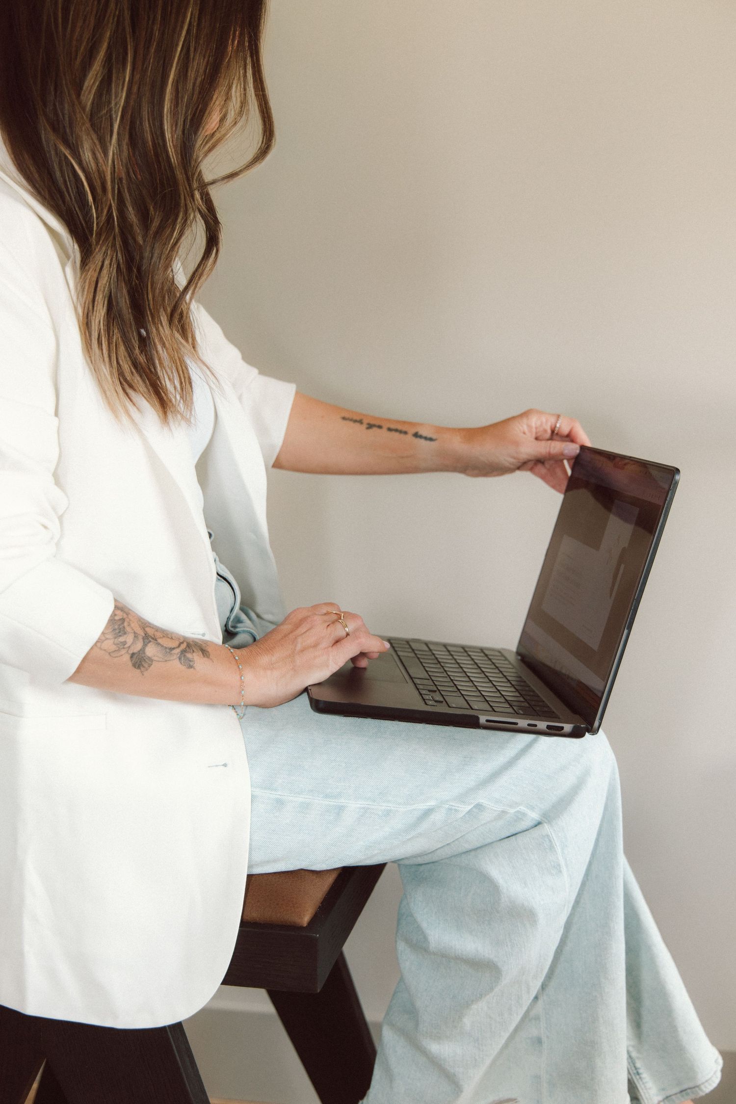 Woman with tattoos in white blazer and blue pants sitting on chair, opening a laptop computer.