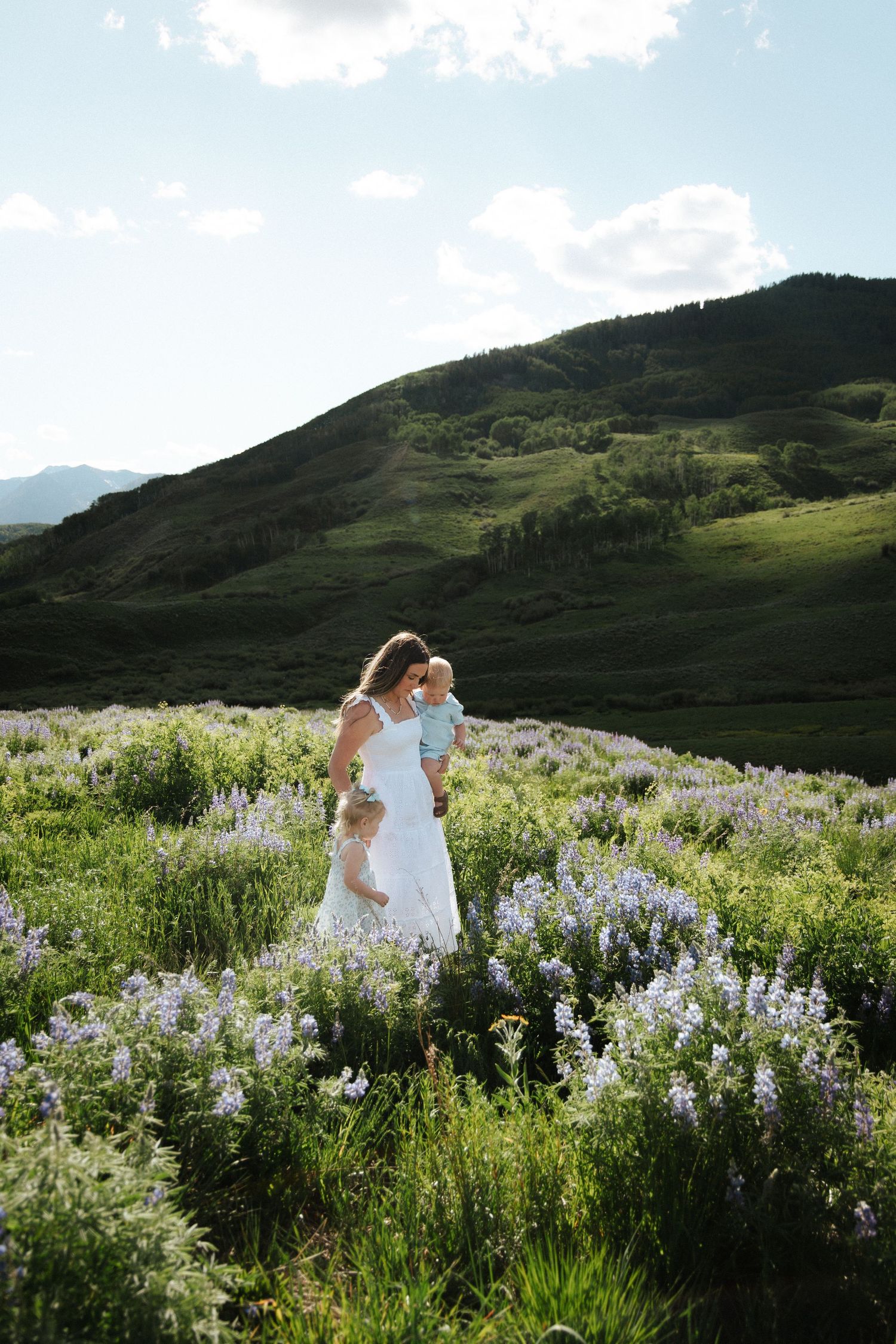 A mother in a white dress holds two young children in a blooming wildflower meadow with green hills in the background.