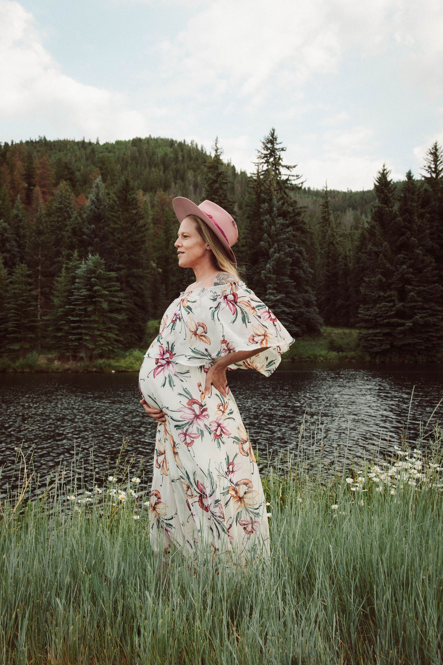 Floral dress captured in tall grass by mountain lake with evergreen trees.