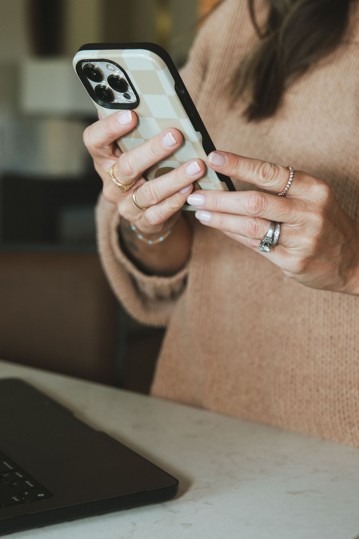 Woman in beige sweater using iPhone with checkered case, wearing rings, seated at marble desk with laptop.