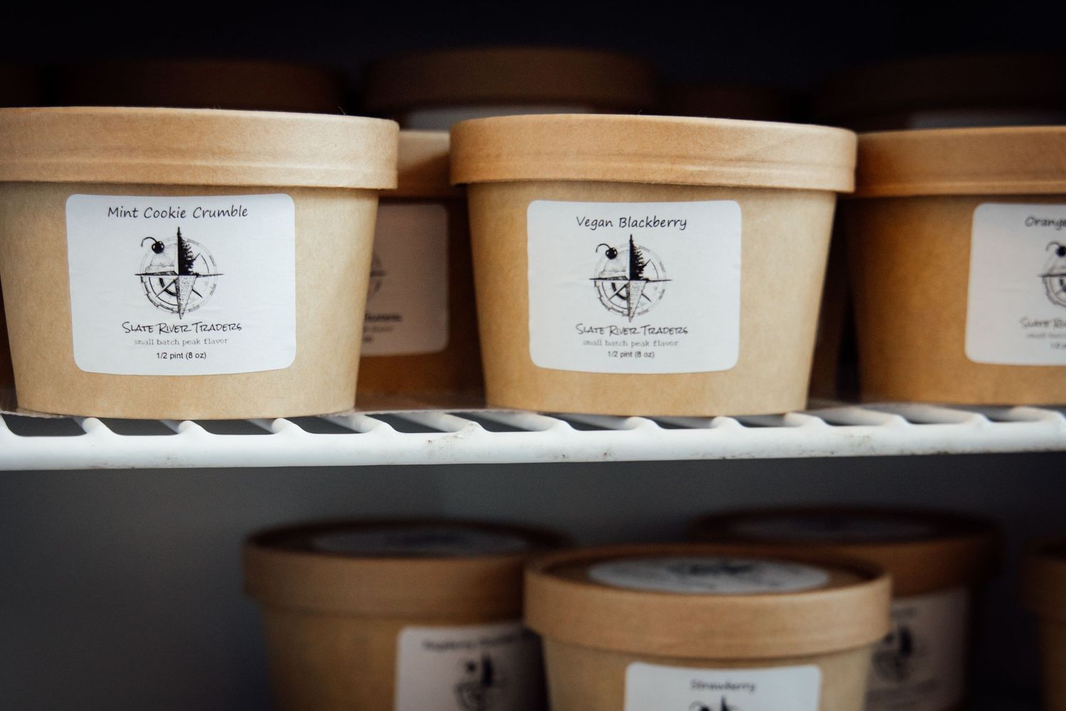 Kraft paper cups of artisan ice cream flavors on a freezer shelf, including Mint Cookie Crumble and Vegan Blackberry.