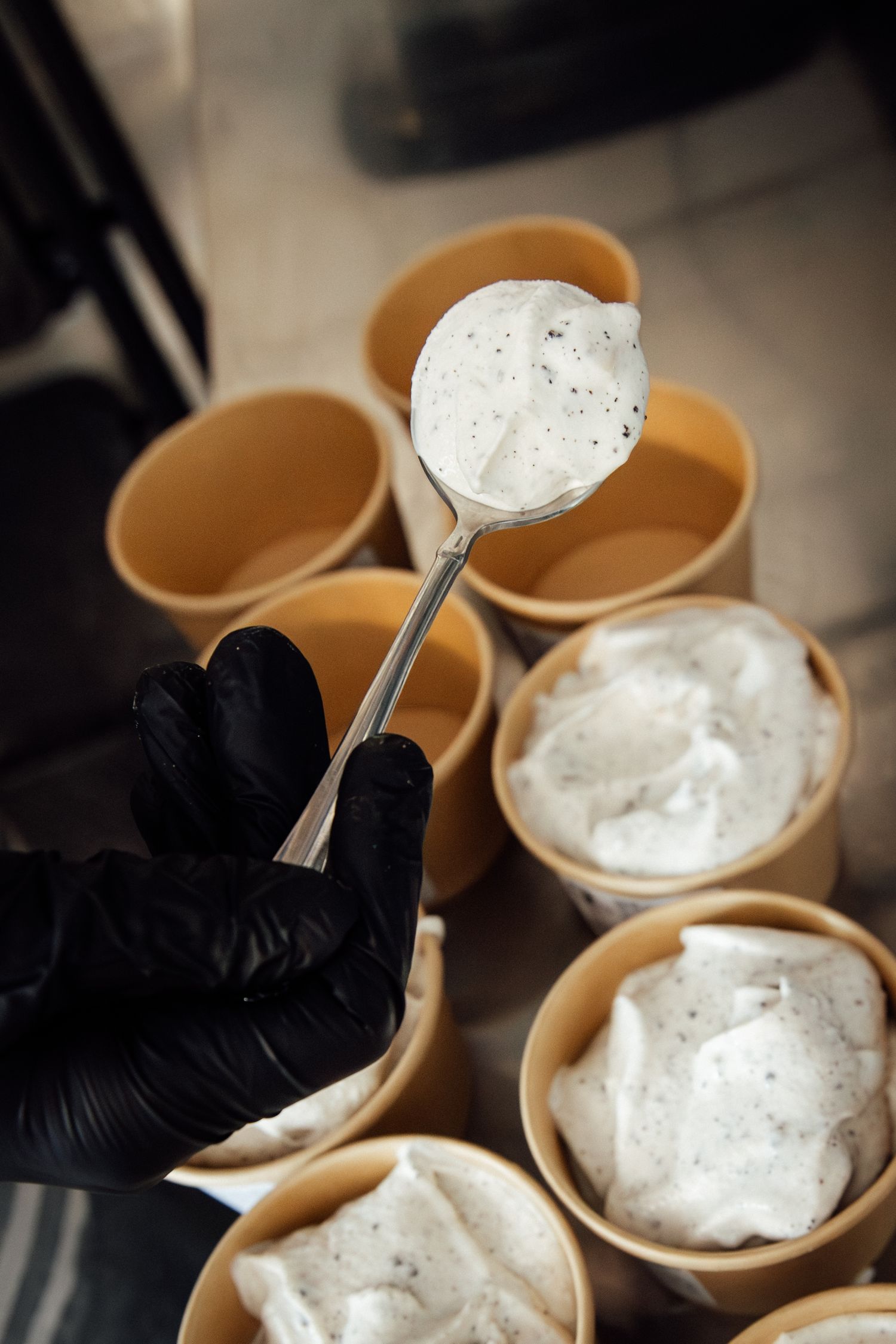Gloved hand holding a spoon of vanilla bean ice cream over paper cups filled with scoops at an ice cream shop.