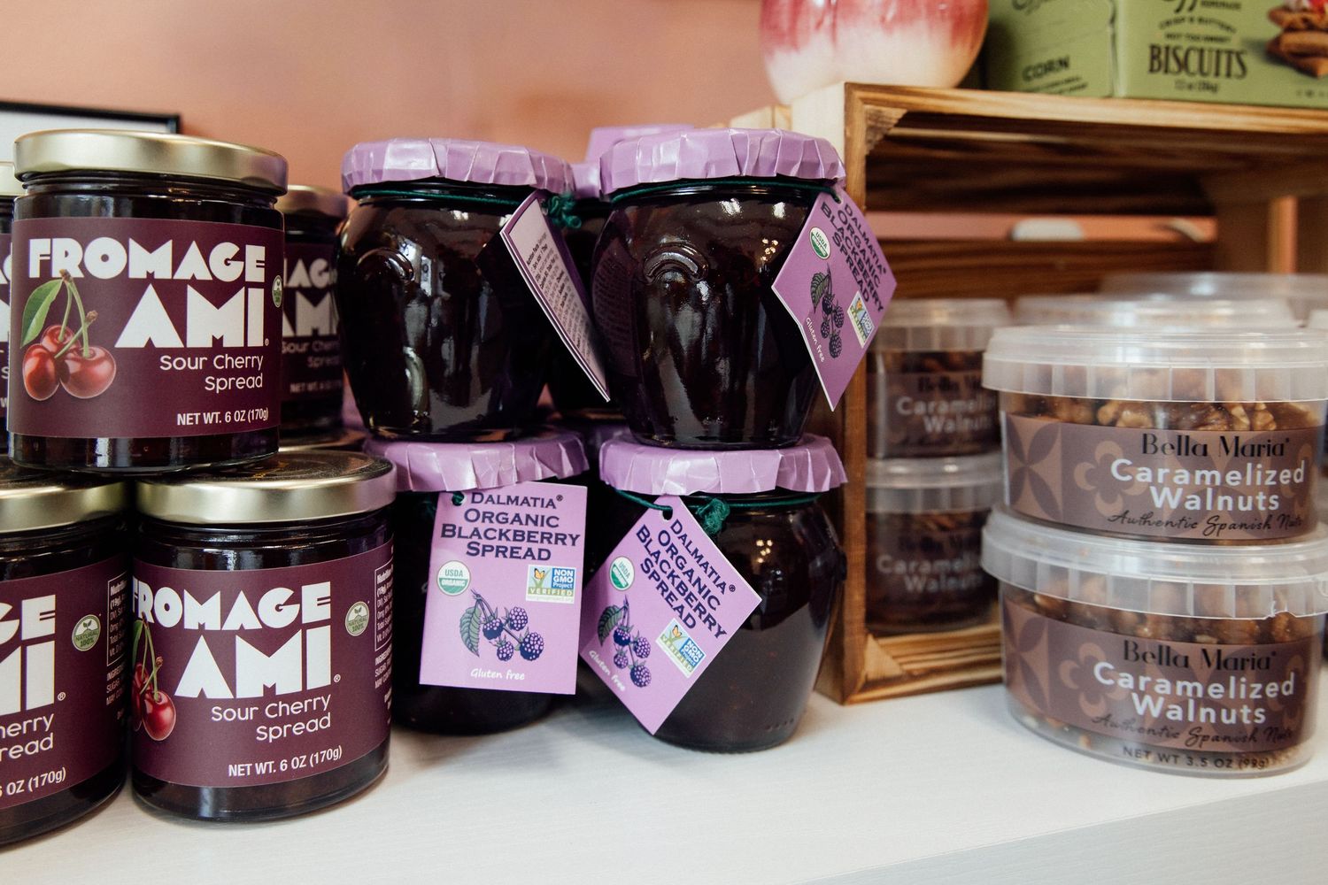 Fromage Ami sour cherry spread jars and caramelized walnuts displayed on a white shelf in a gourmet shop.