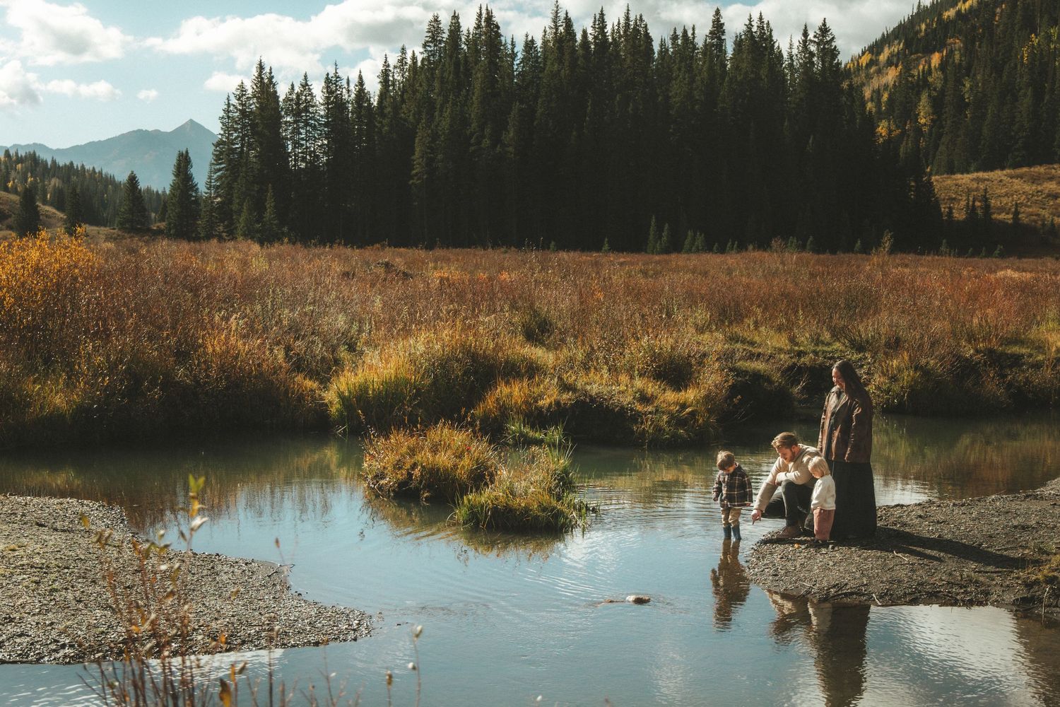 Family with children playing at the edge of a calm mountain stream surrounded by golden autumn grasses and evergreen forest.