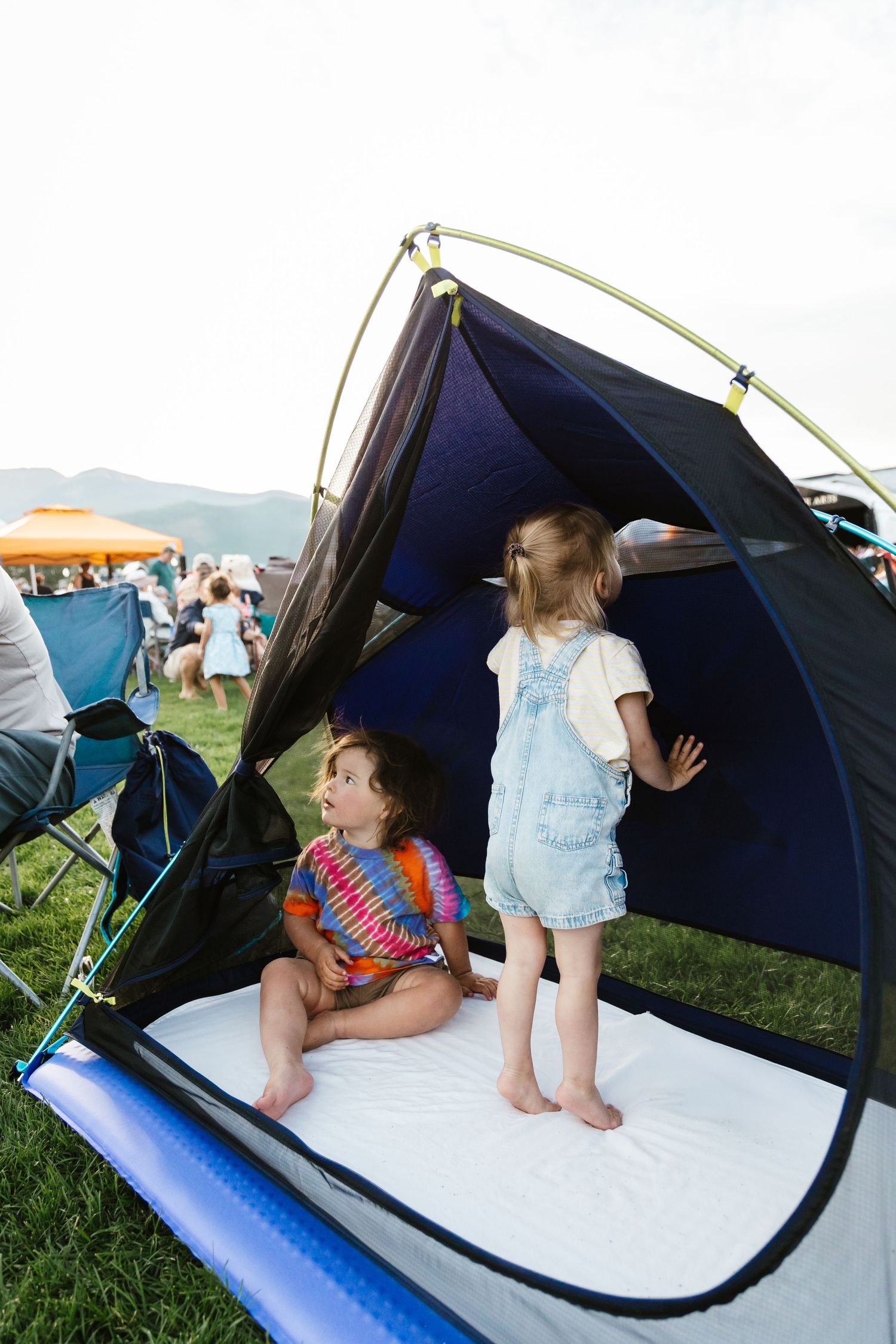 Small blue camping tent set up on grass at outdoor gathering with mountains visible in background.