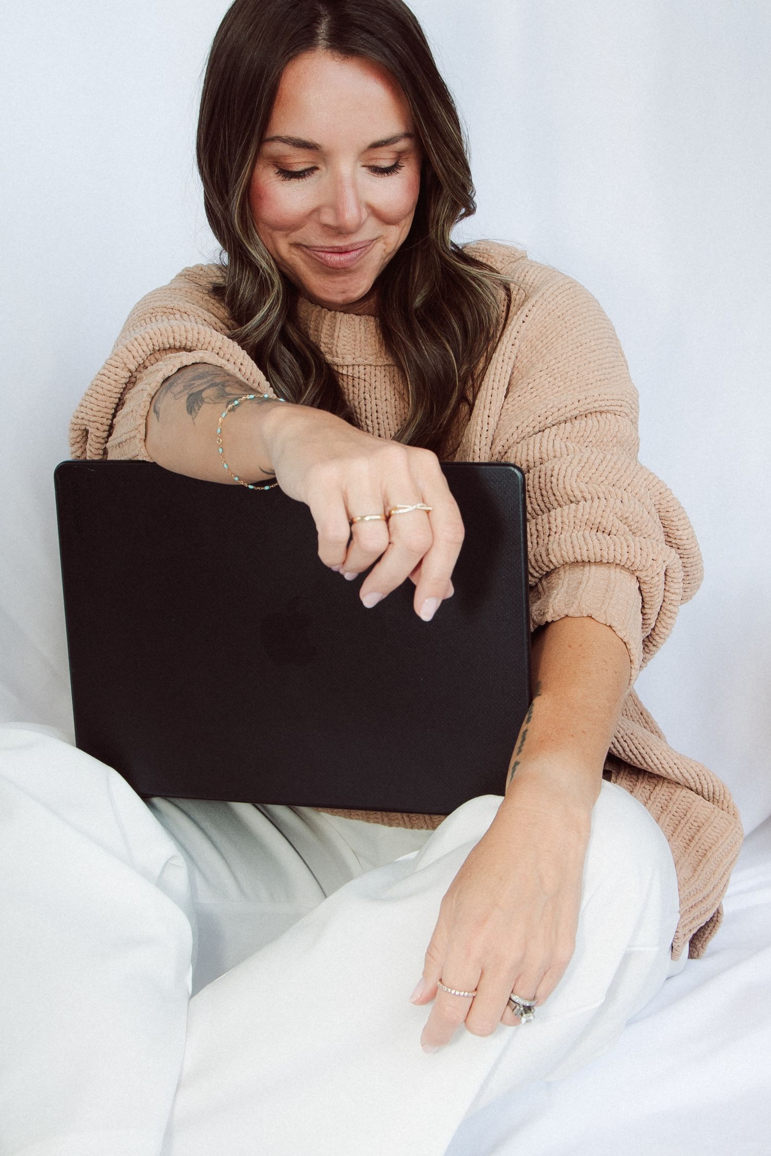Woman in beige sweater smiling while using a black laptop, sitting on white bedding.