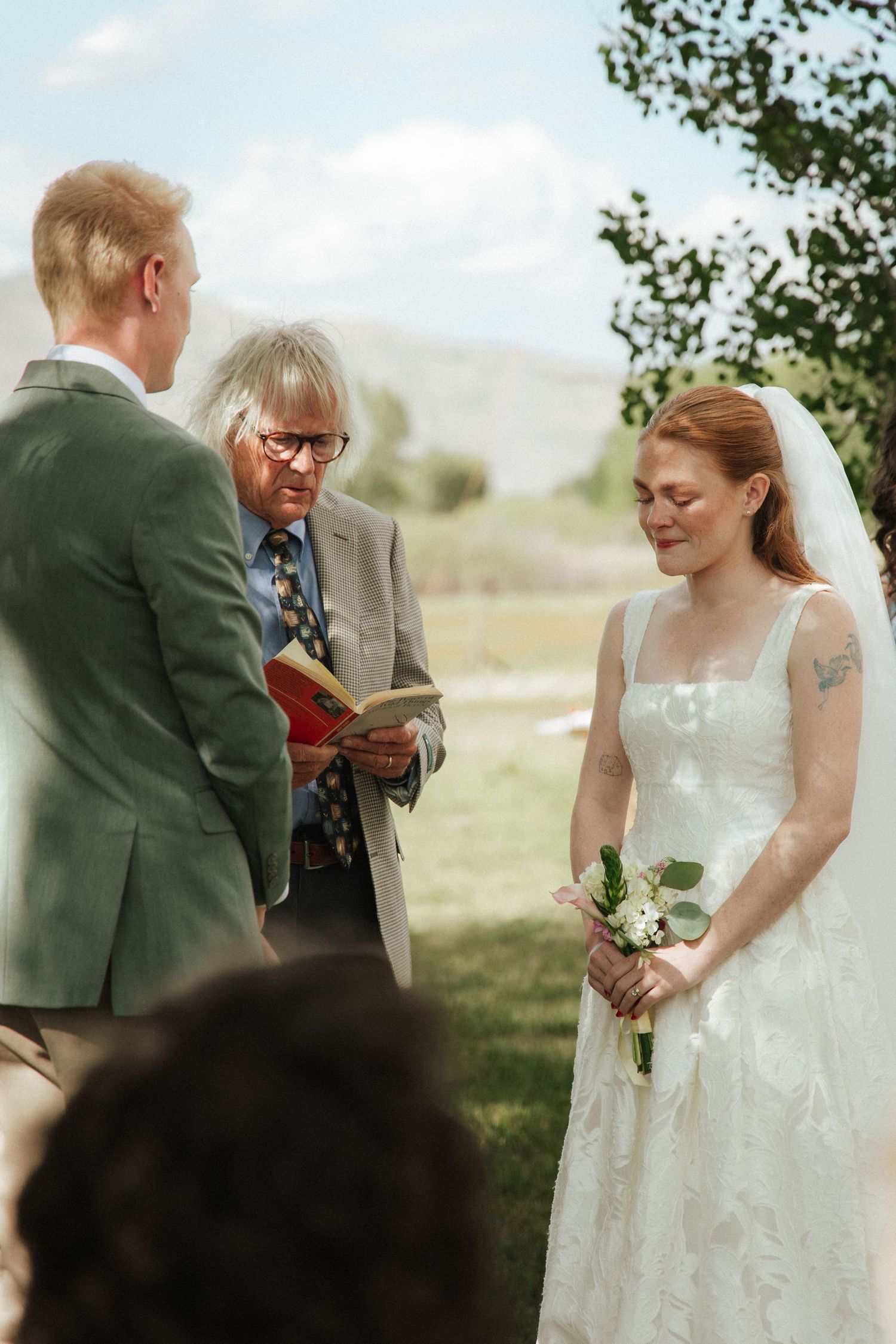 Outdoor wedding ceremony with bride in white lace gown holding bouquet, groom in green suit, and officiant reading vows.