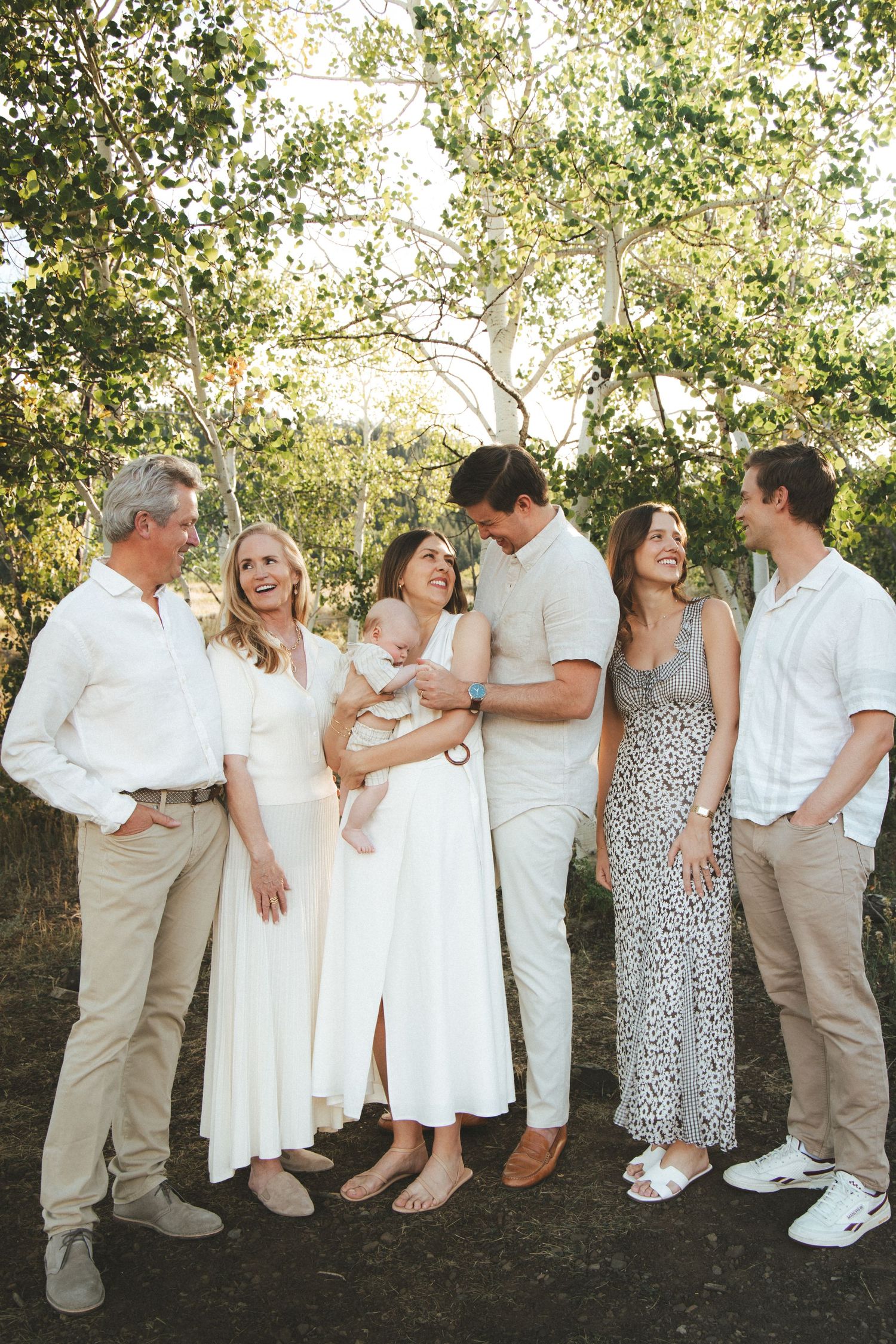 A joyful multigenerational family in white and neutral tones laughing together outdoors among lush green trees.