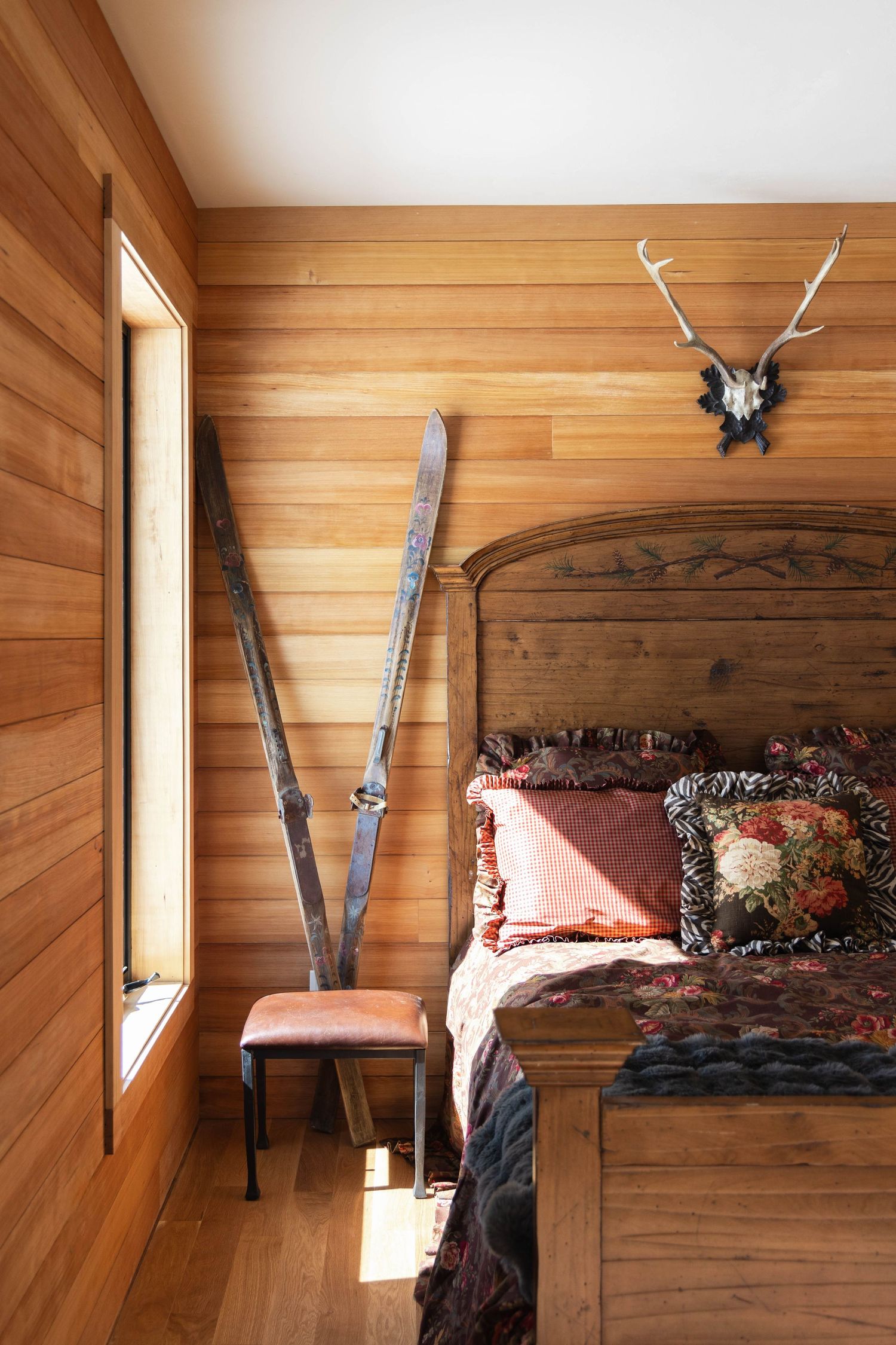 Rustic wood-paneled bedroom with vintage skis, antler mount, ornate wooden bed frame, and rich burgundy bedding.