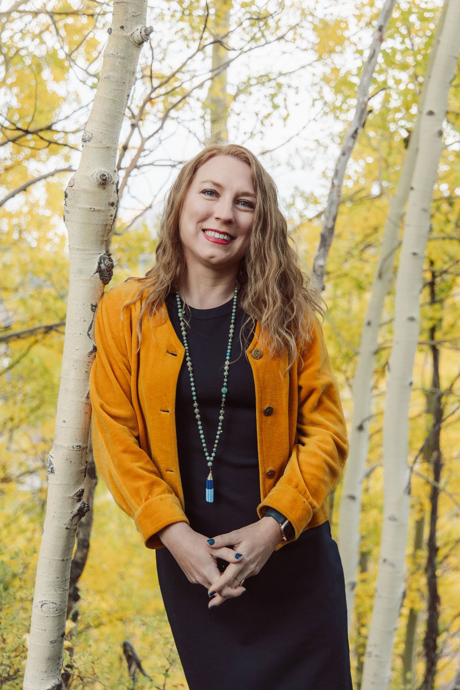 A person wearing a mustard yellow jacket and black dress poses among aspen trees with golden autumn foliage.
