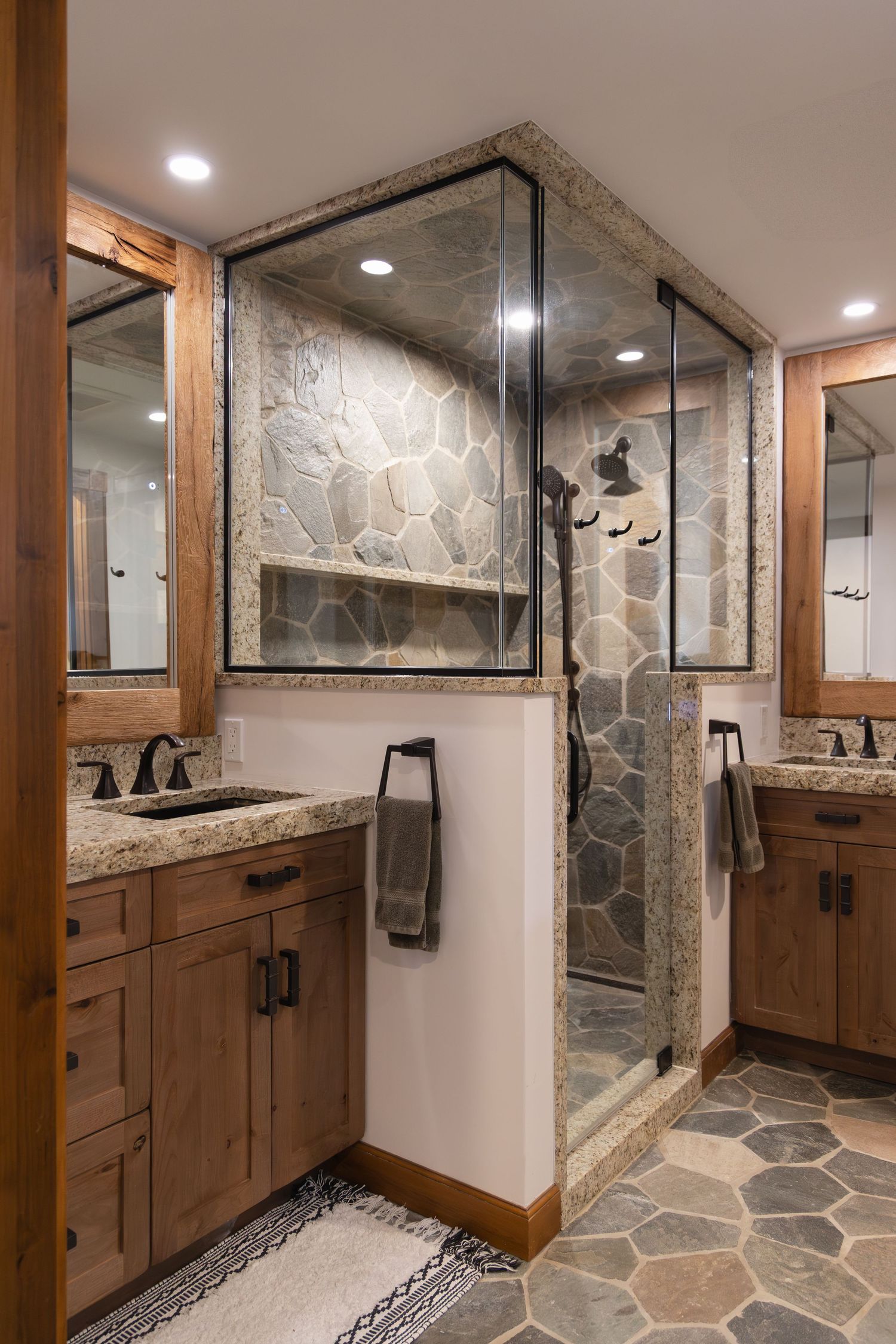 Rustic bathroom with stone tile walk-in shower, wood vanities, granite countertops, and hexagonal slate floor tiles.
