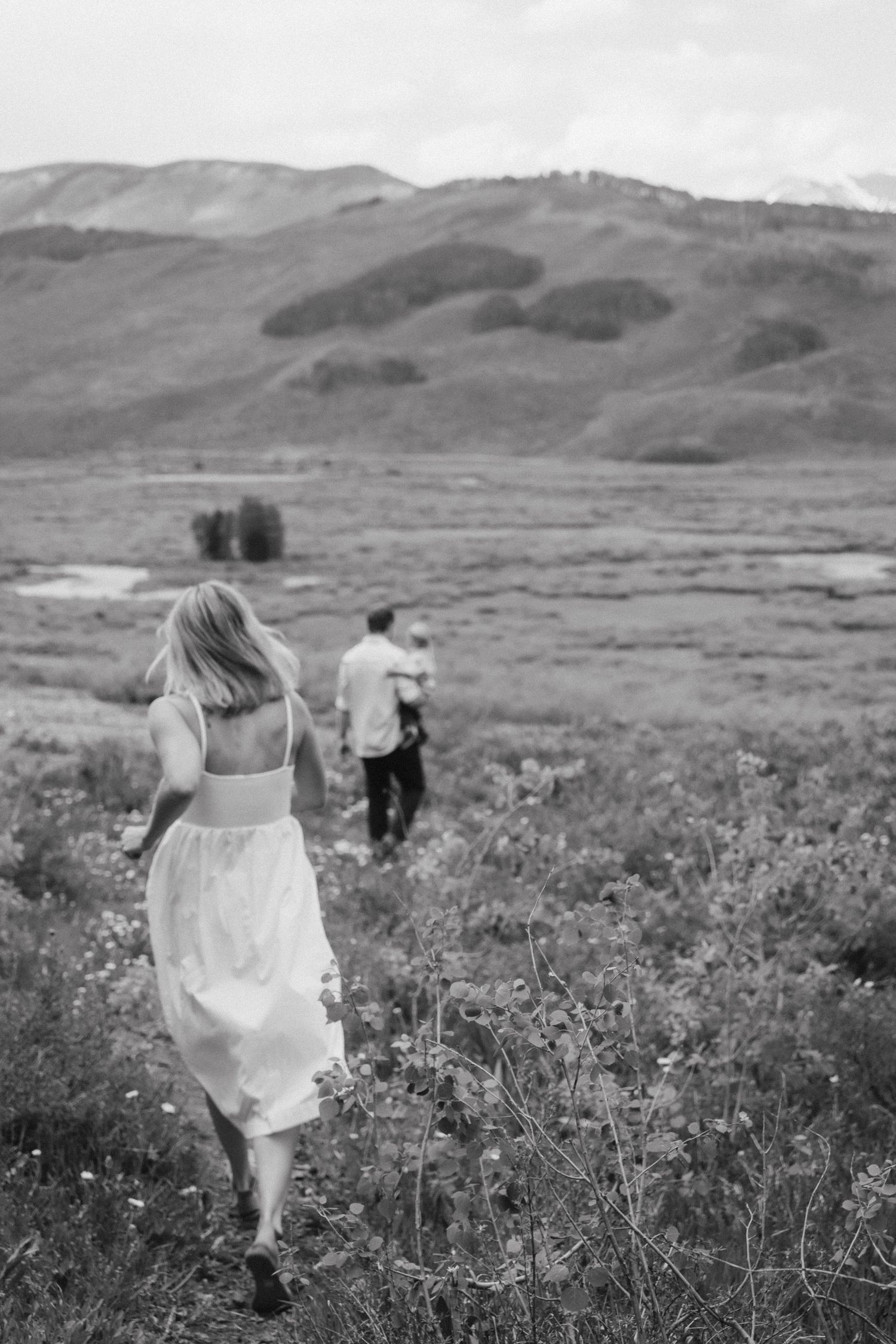 Black and white photo of couple running through wildflower meadow with mountains in background.