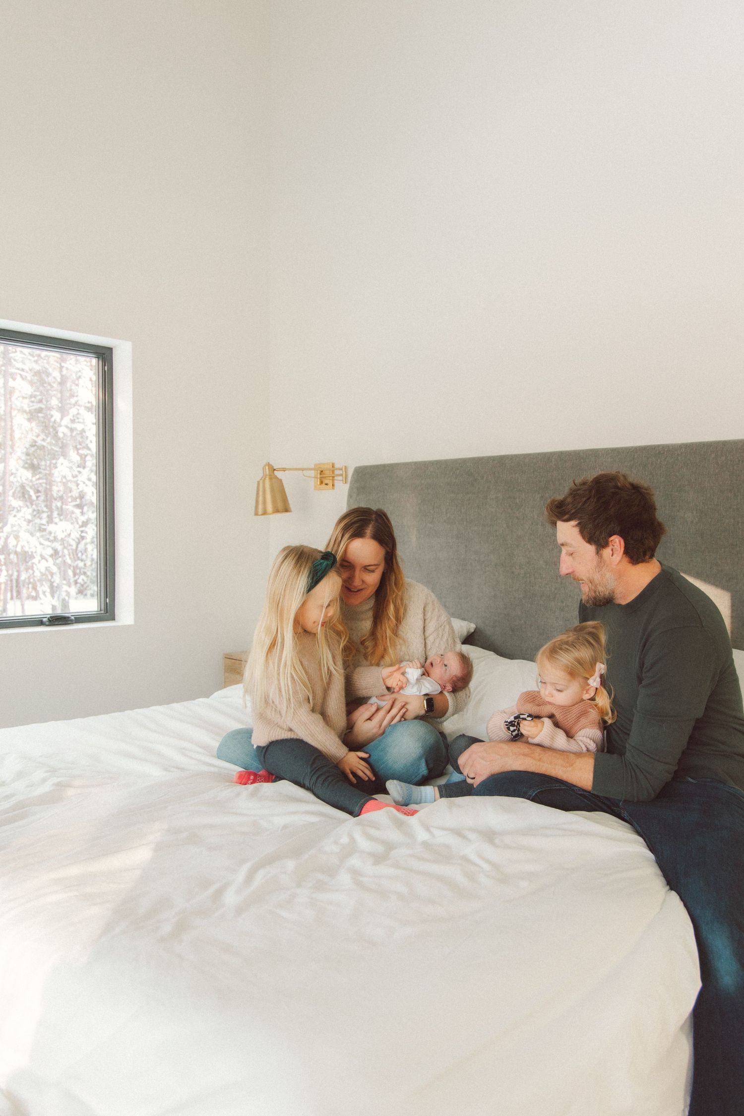 A family shares tender moments together while relaxing on a white bed in a bright, minimalist bedroom.