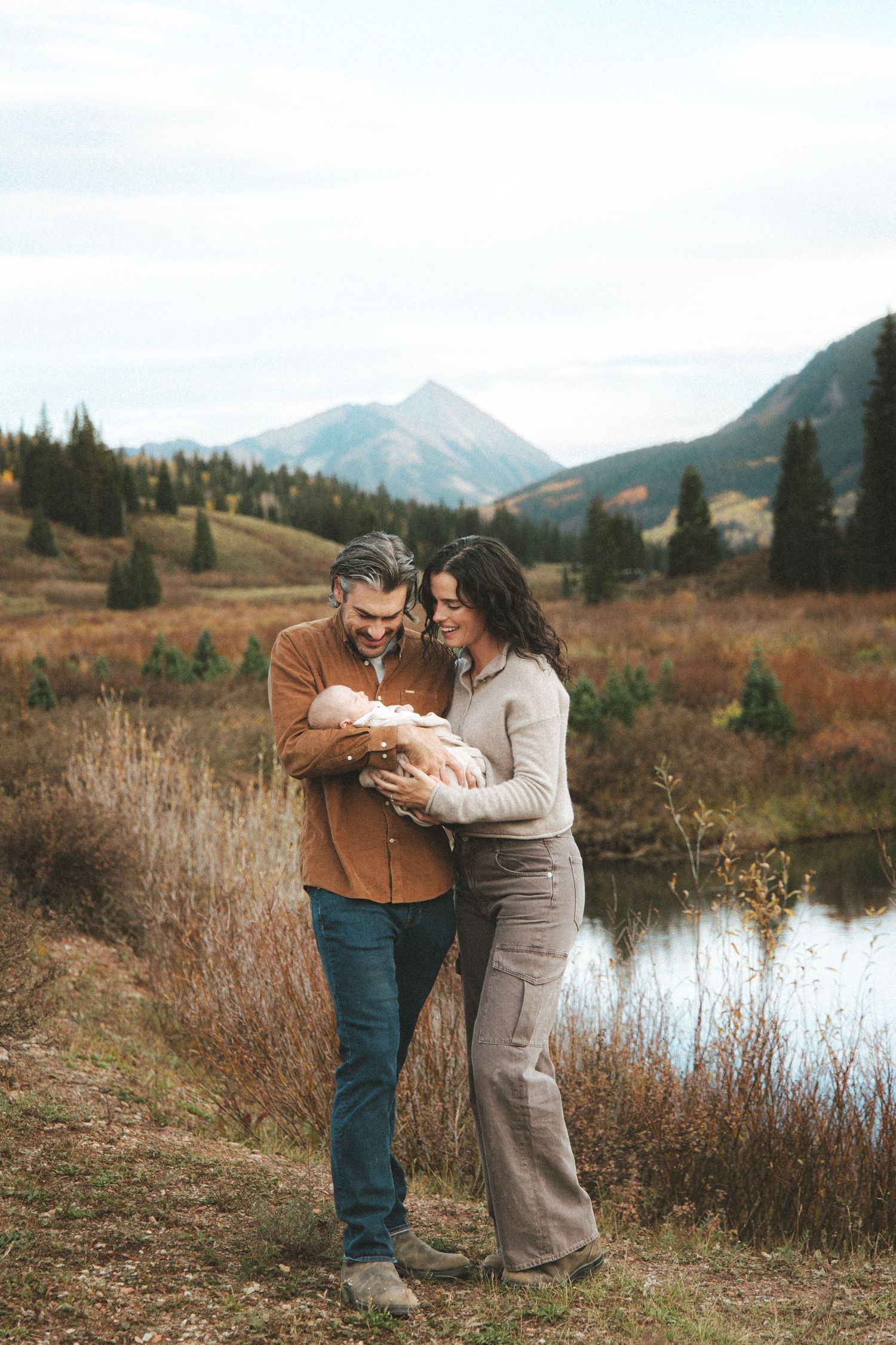 A couple smiles at their newborn baby outdoors near a mountain lake in autumn.