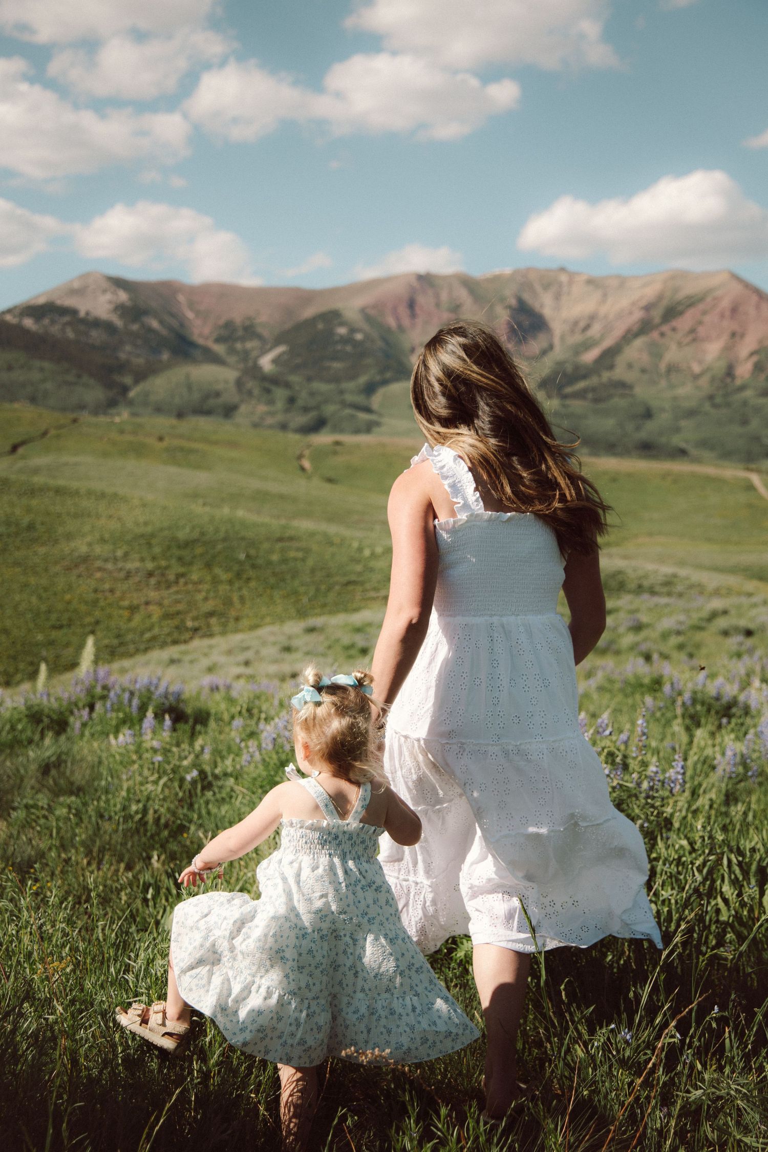 Mother and daughter in white dresses walking through green meadow with mountain backdrop.