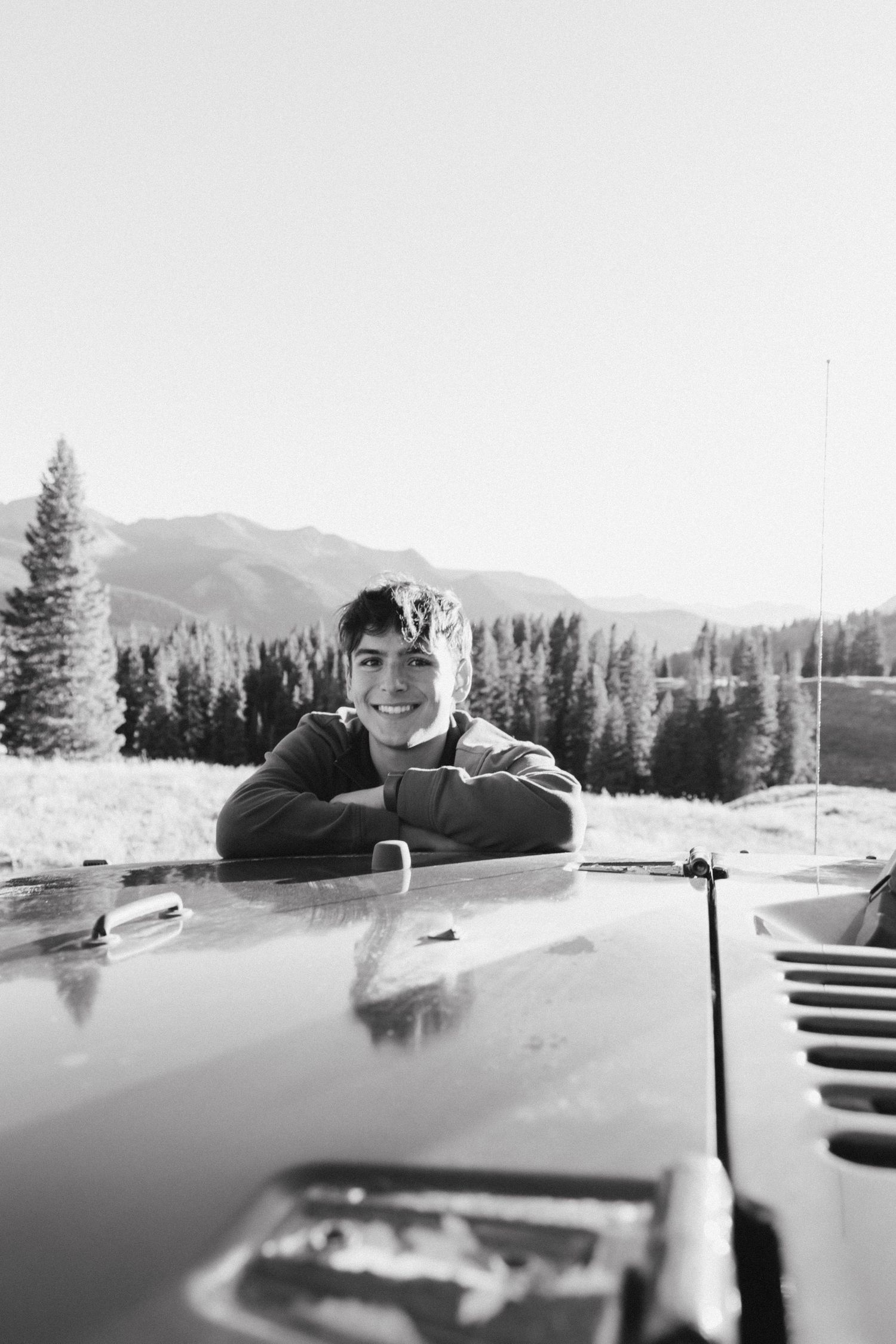 Black and white portrait of someone smiling through a car window in a mountain setting.