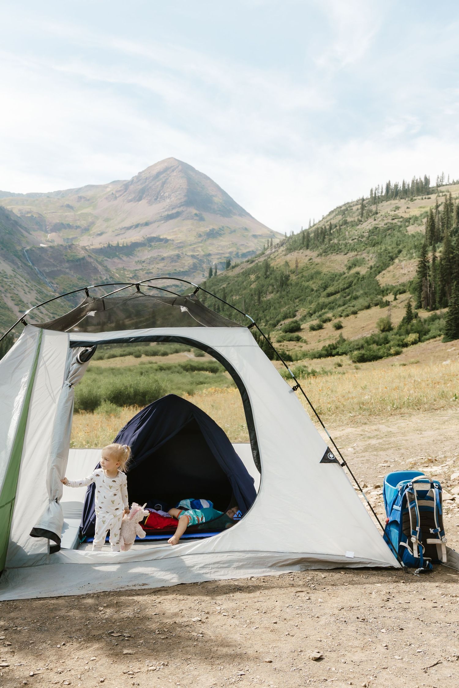 A sequence of four camping tents set up in a mountainous valley with rugged peaks and wilderness terrain in the background.