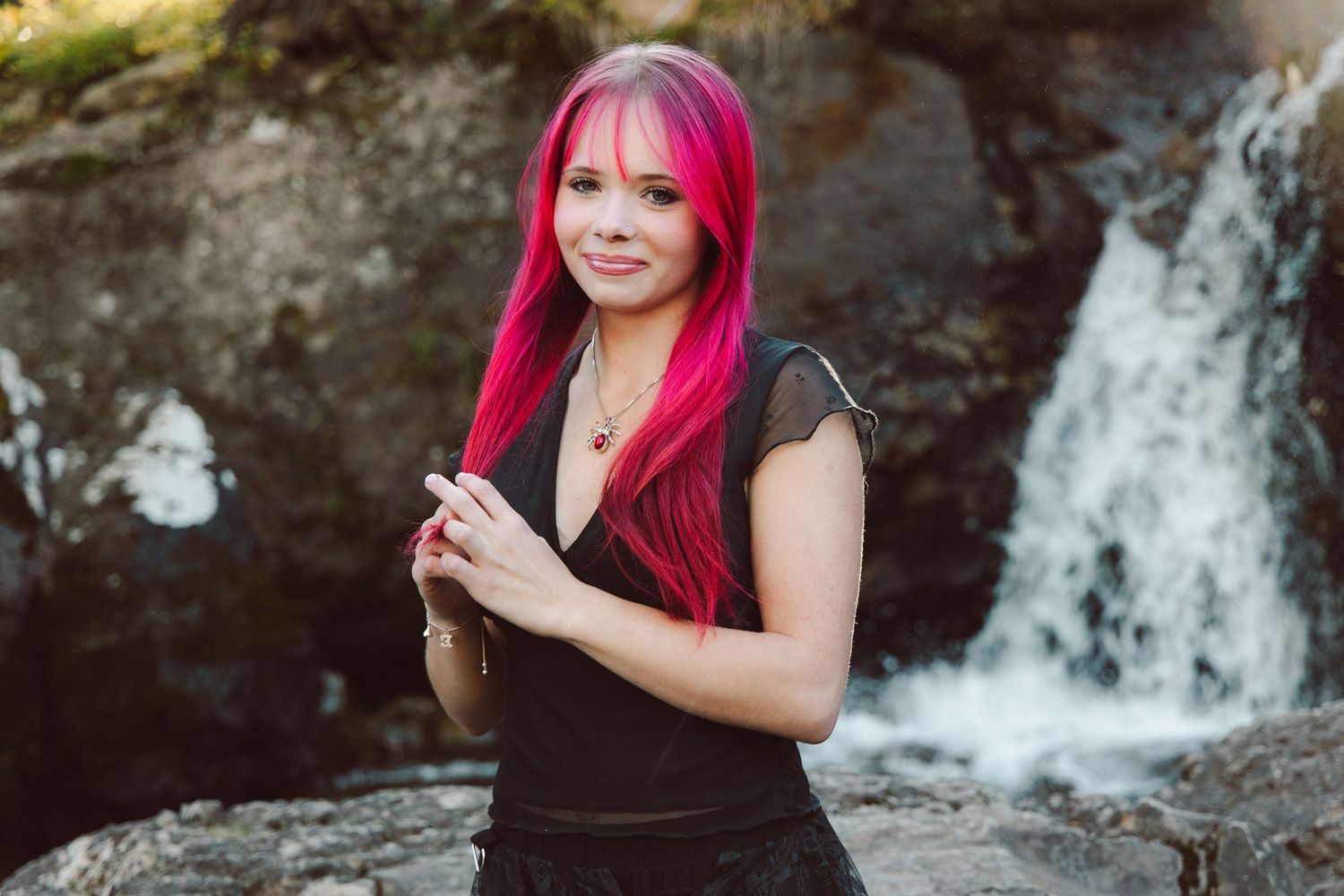 Woman with vibrant pink hair poses gracefully near a waterfall, wearing a black top in a serene outdoor setting.
