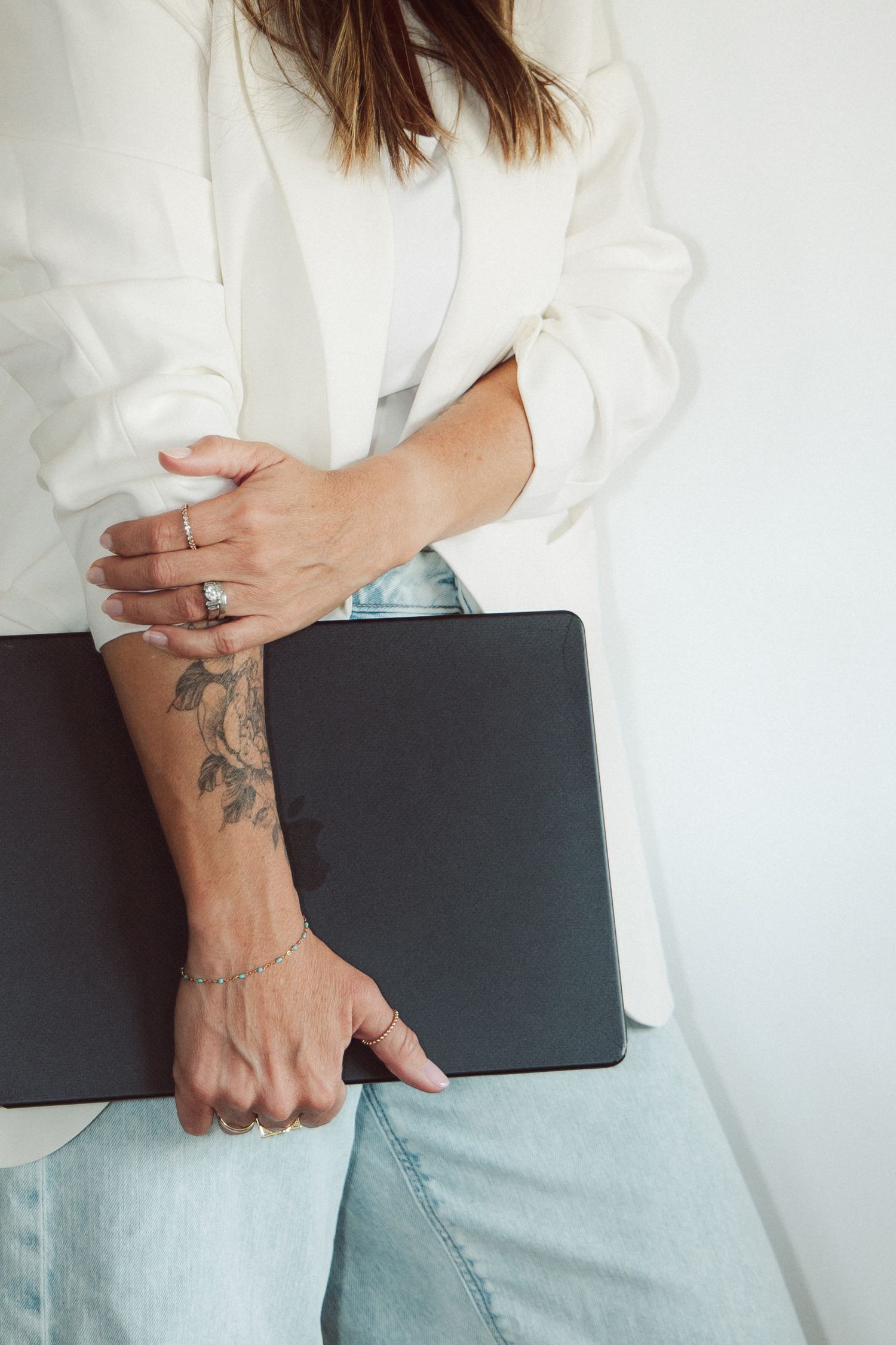 Woman in white blazer and jeans holding a black portfolio, showcasing a floral forearm tattoo and rings.
