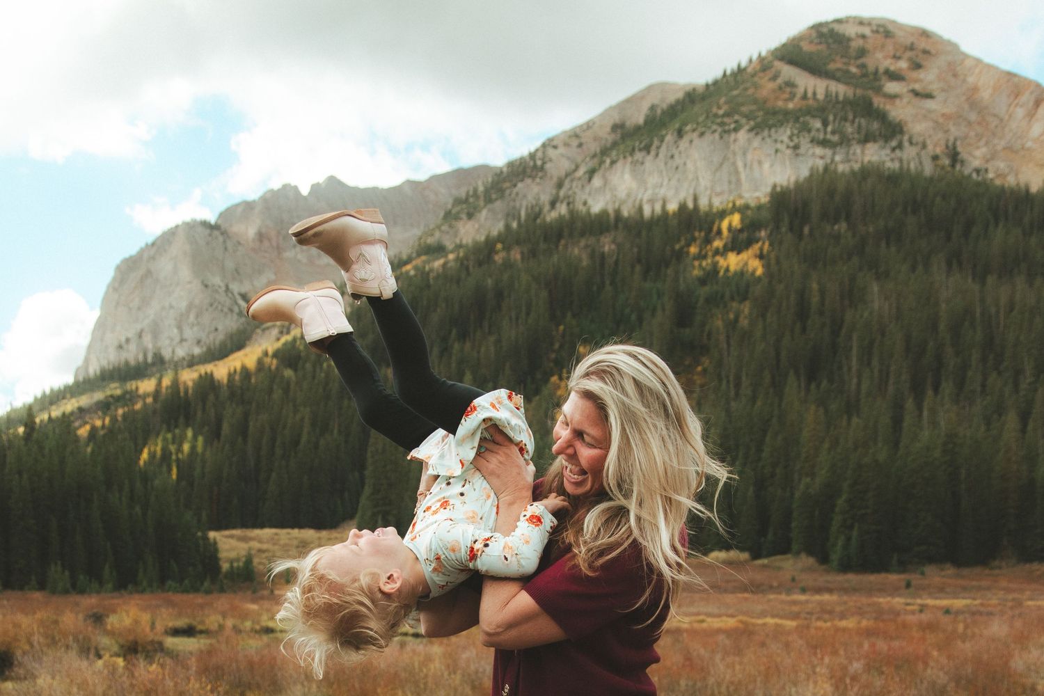 A joyful mother playfully lifting her toddler outdoors with stunning mountain scenery in the background.