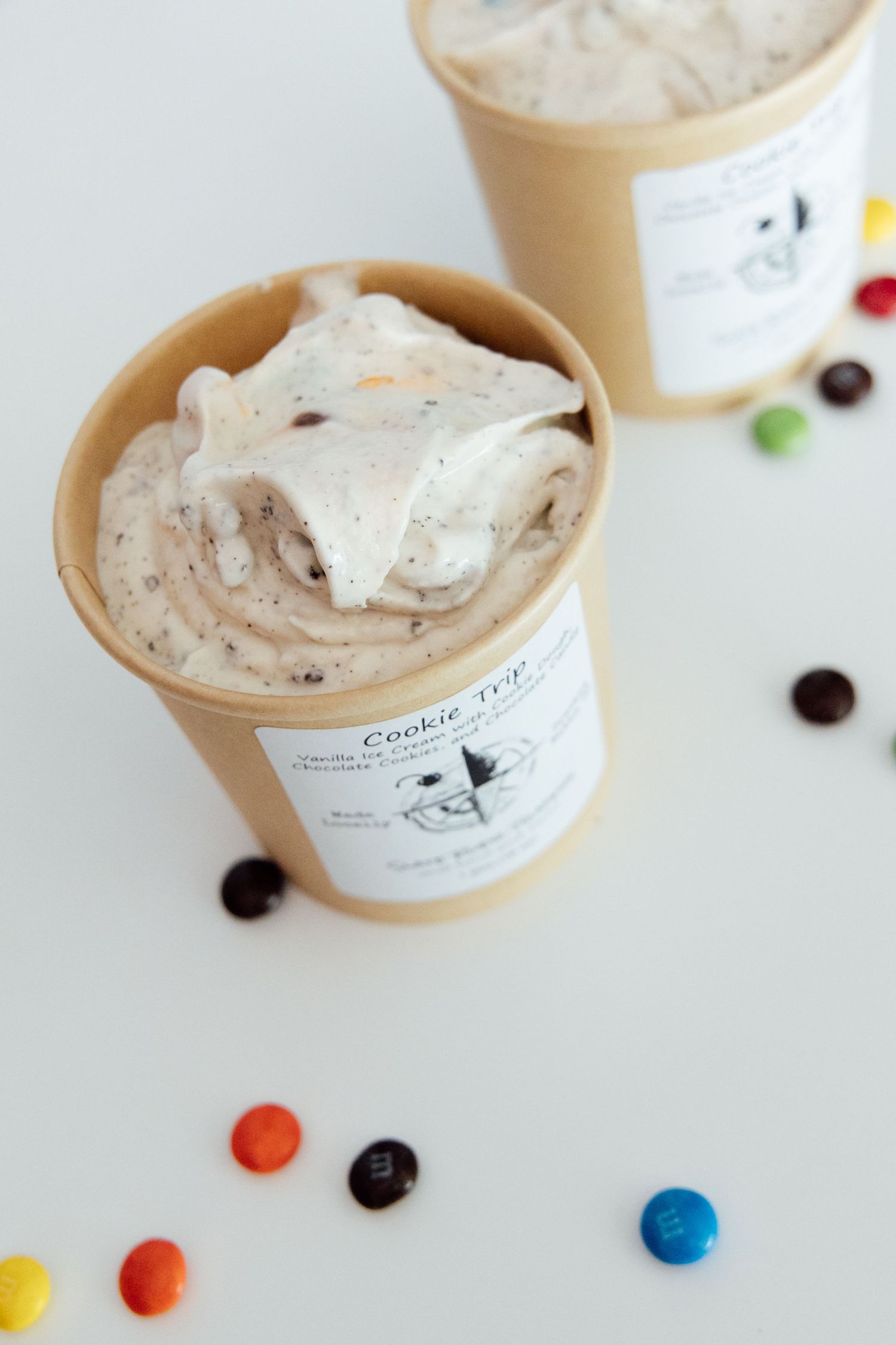 Two paper cups of Cookie Top ice cream with M&M candies scattered on a white surface.