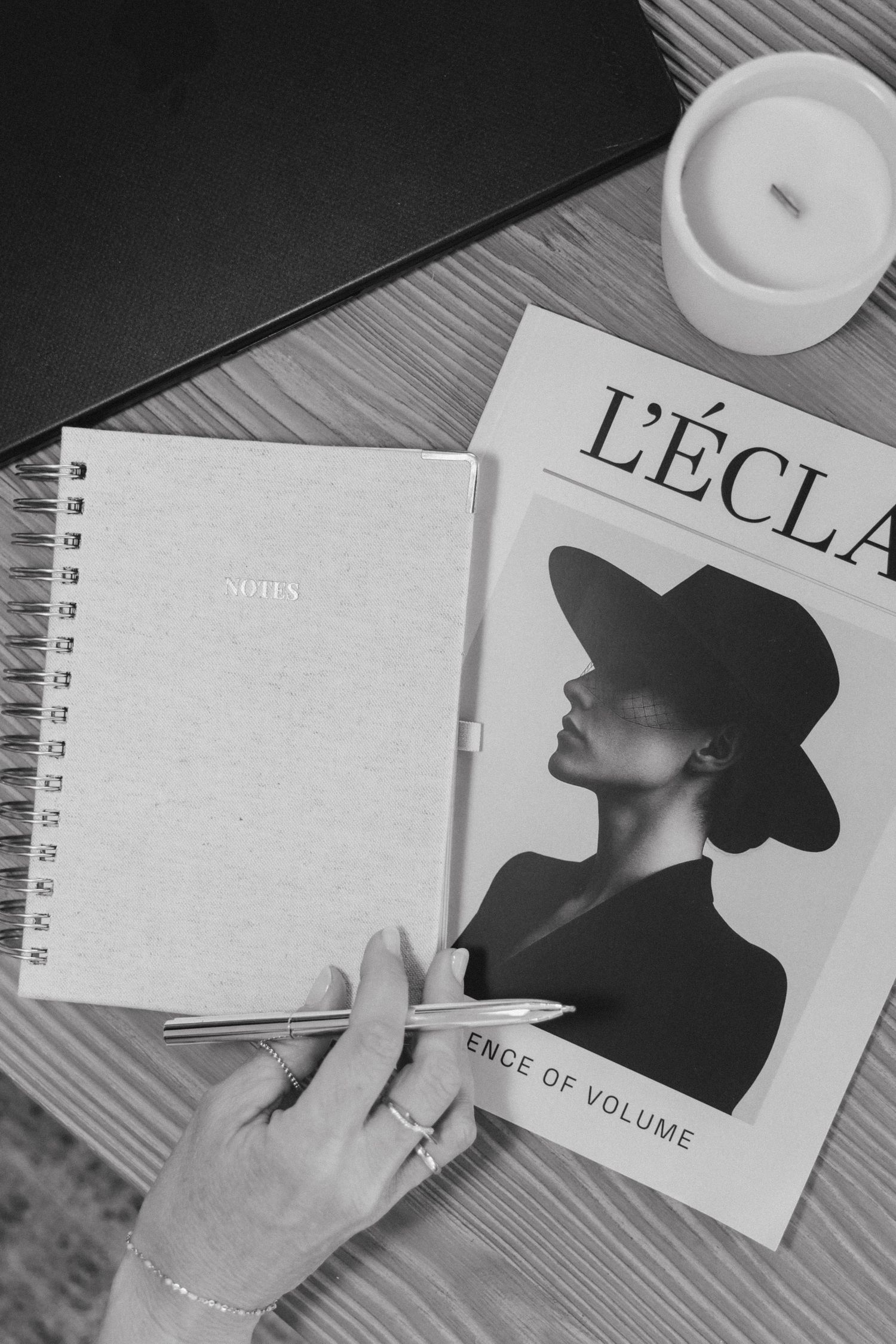 Black and white flatlay of a spiral notebook, L'Eclat magazine, pen, and candle on a wooden desk.