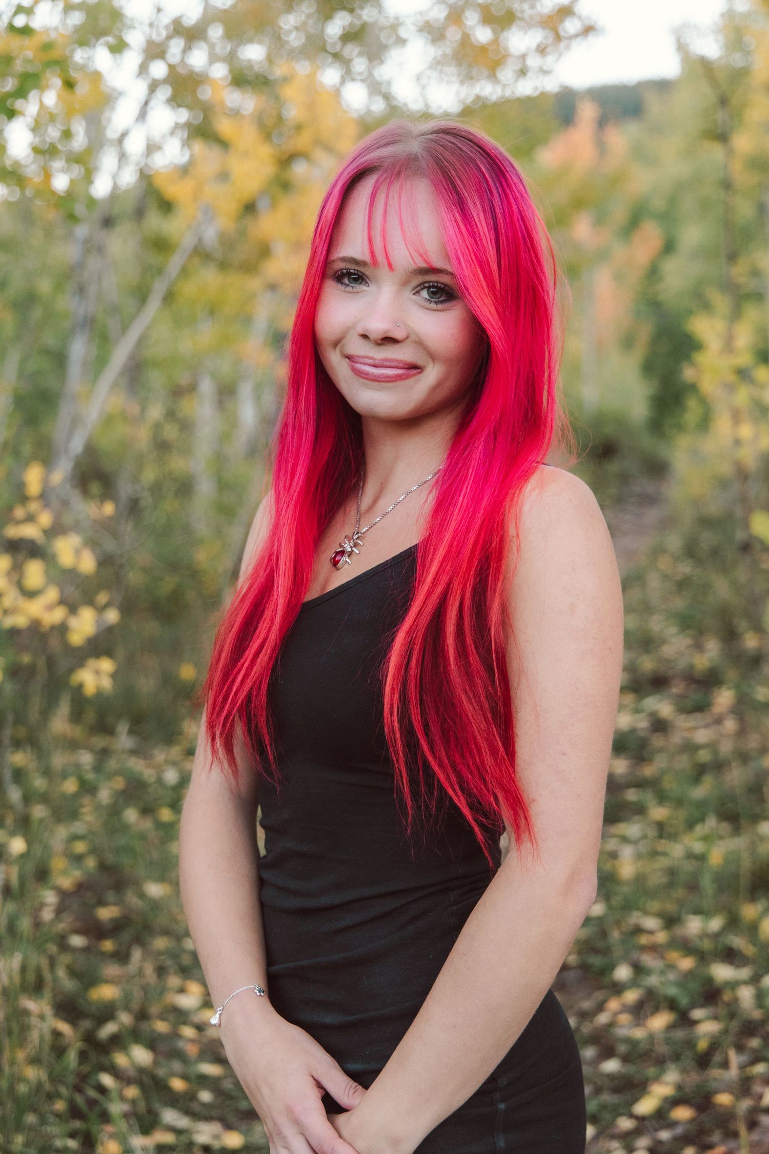 Woman with vibrant pink hair poses outdoors in a black dress against autumn foliage.
