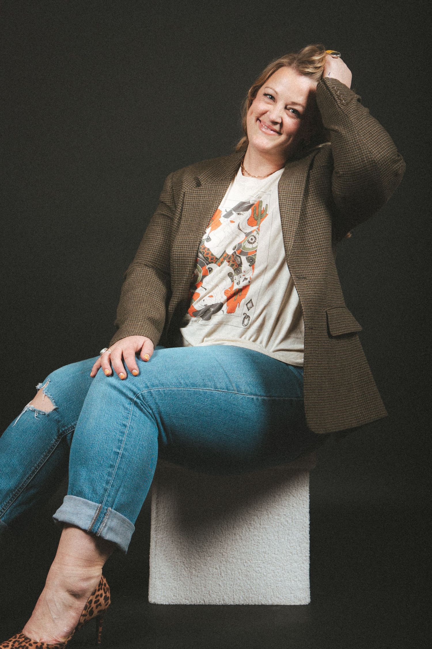 A casual portrait featuring someone wearing a brown blazer, graphic t-shirt and distressed jeans while sitting on a white surface.
